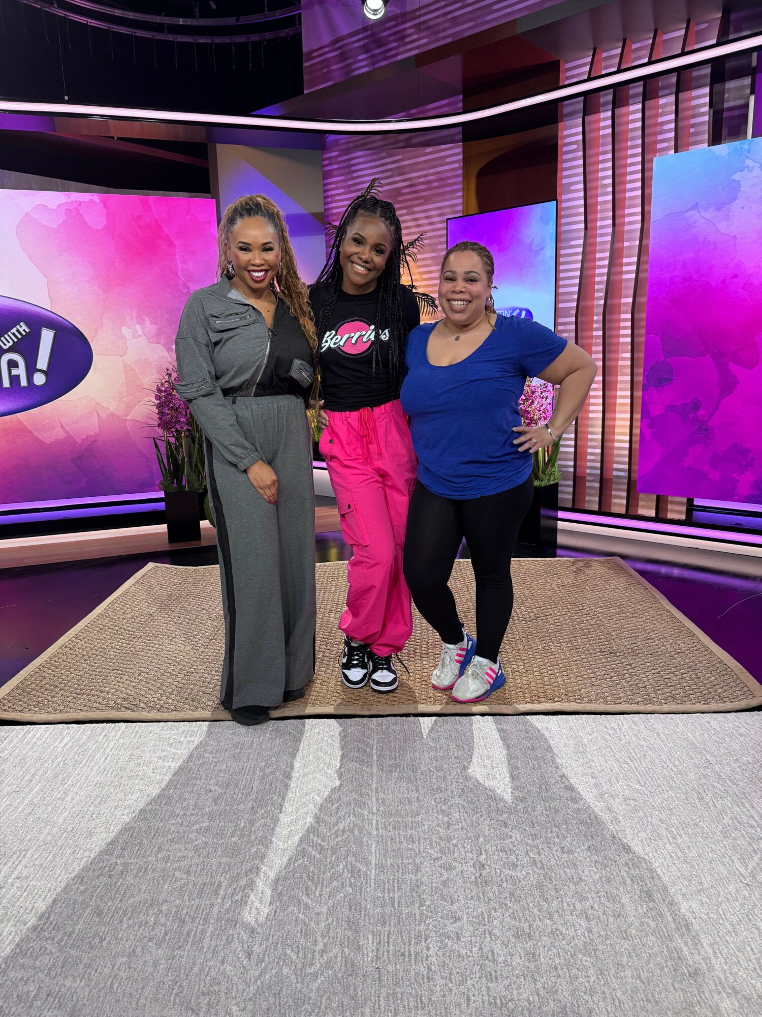 Three women standing on a stage with a pink and purple background, smiling and posing for the photo.