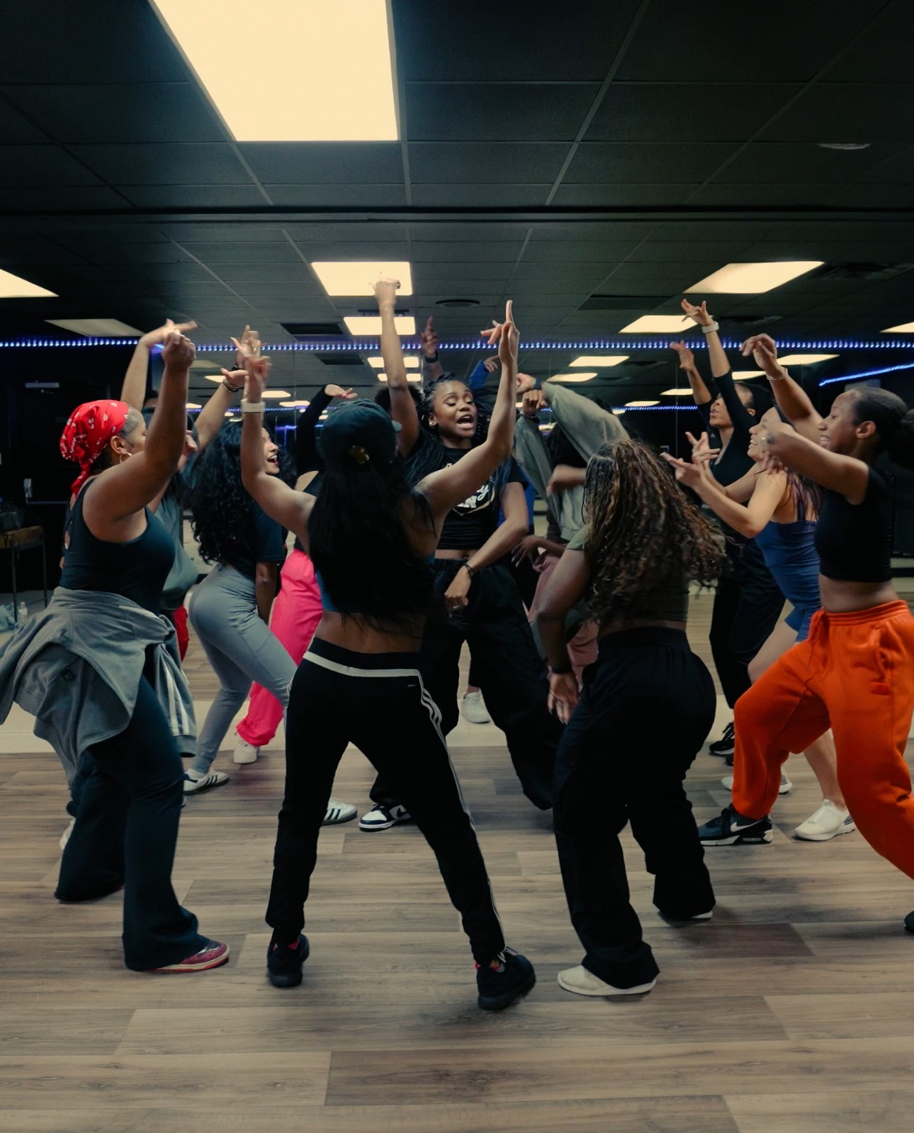 Group of people dancing in a studio with dark ceiling and bright overhead lights, raising hands and moving rhythmically.
