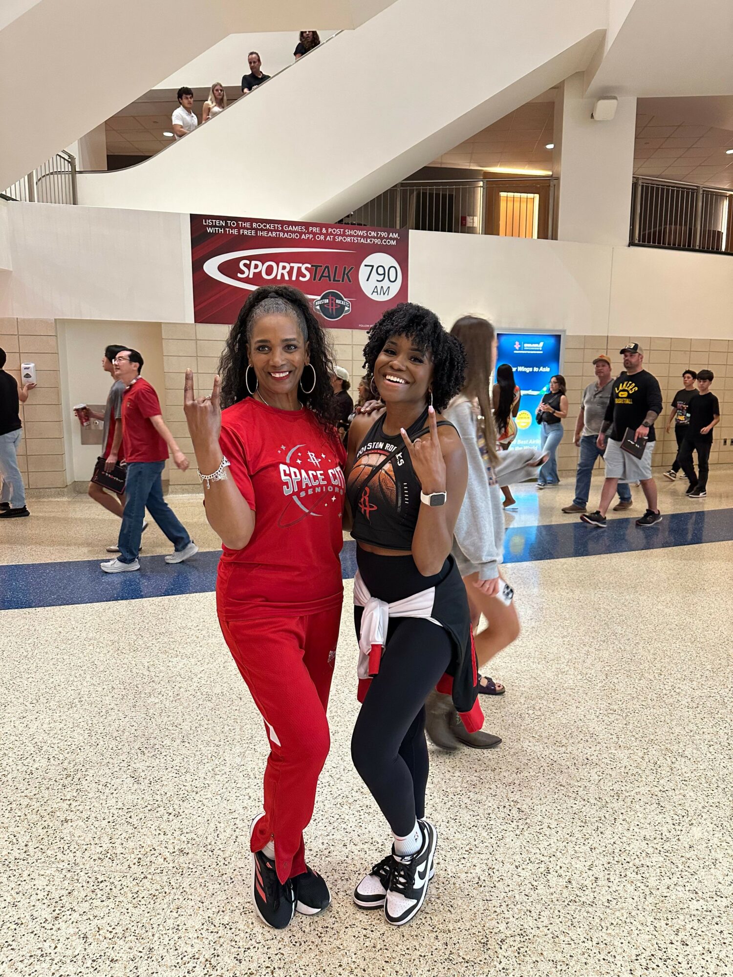 Two women pose together in an indoor public space, one in a red dress and the other in black, with people in the background.