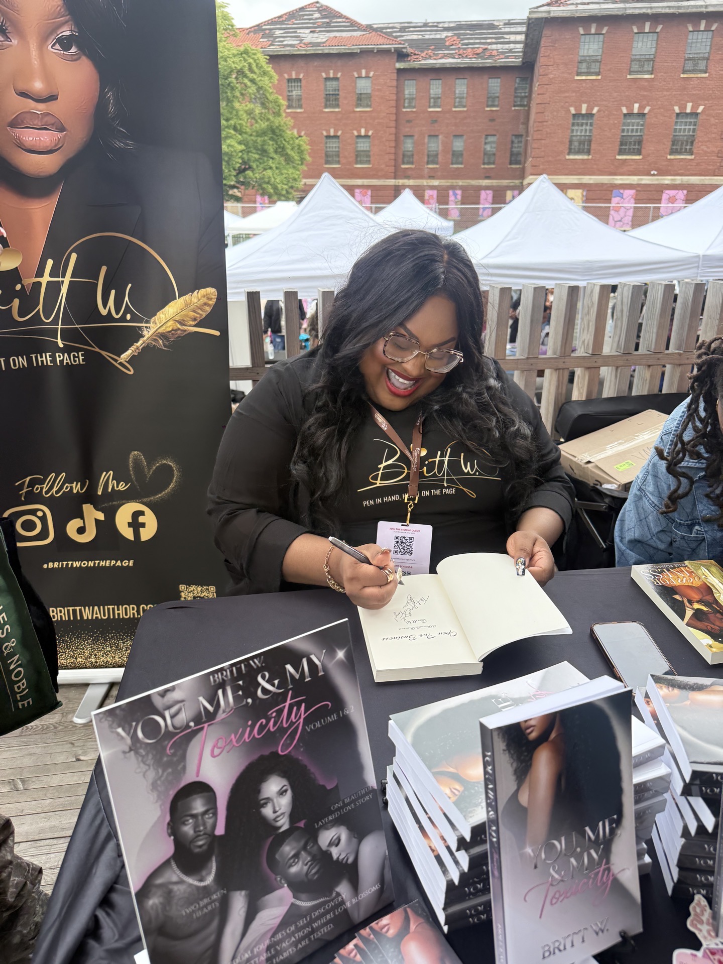 Woman with glasses signing a book at a table outdoors, with promotional materials and tents in background.