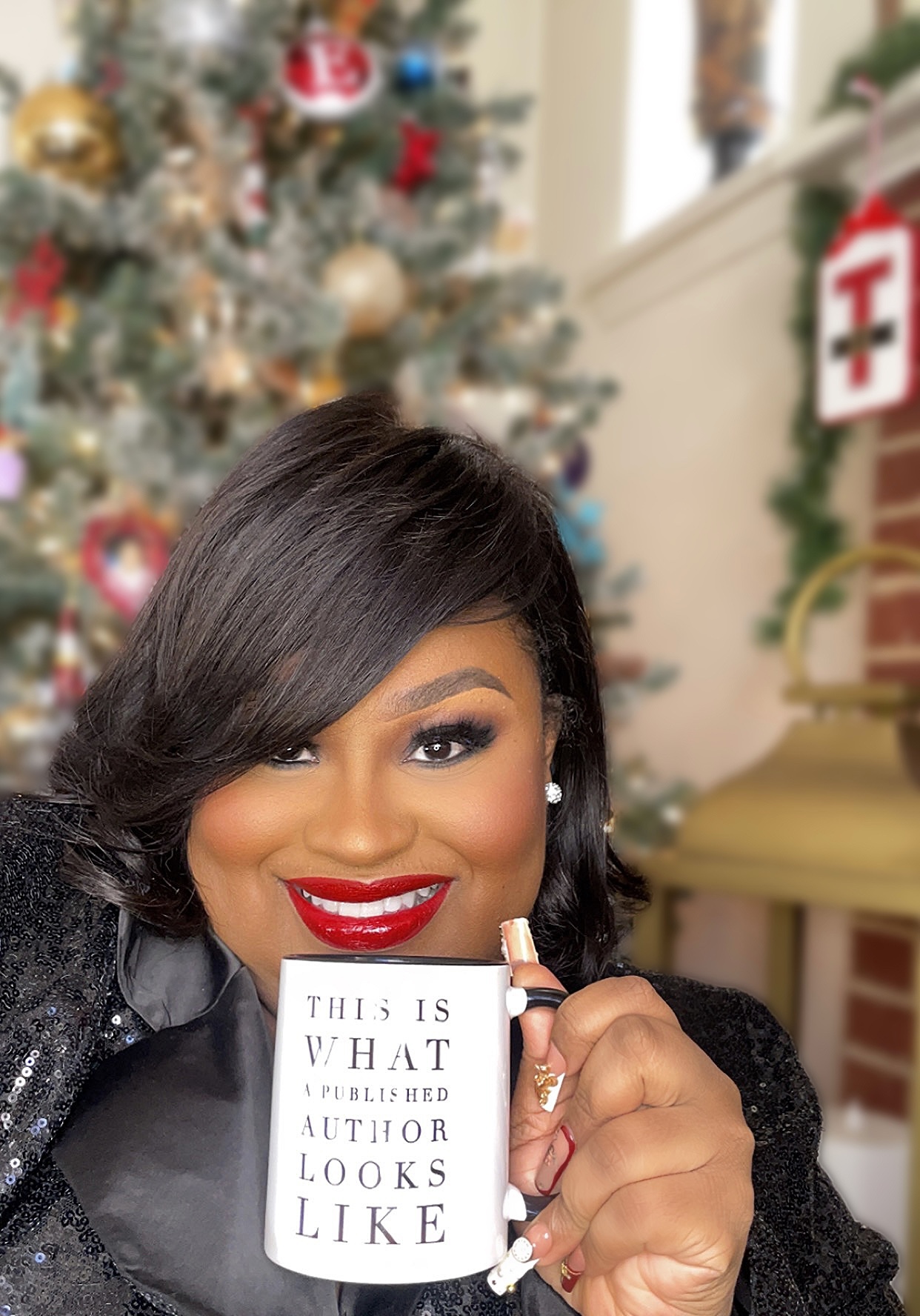 Smiling woman with dark hair and red lipstick holding a white mug with text, decorated Christmas tree in background.