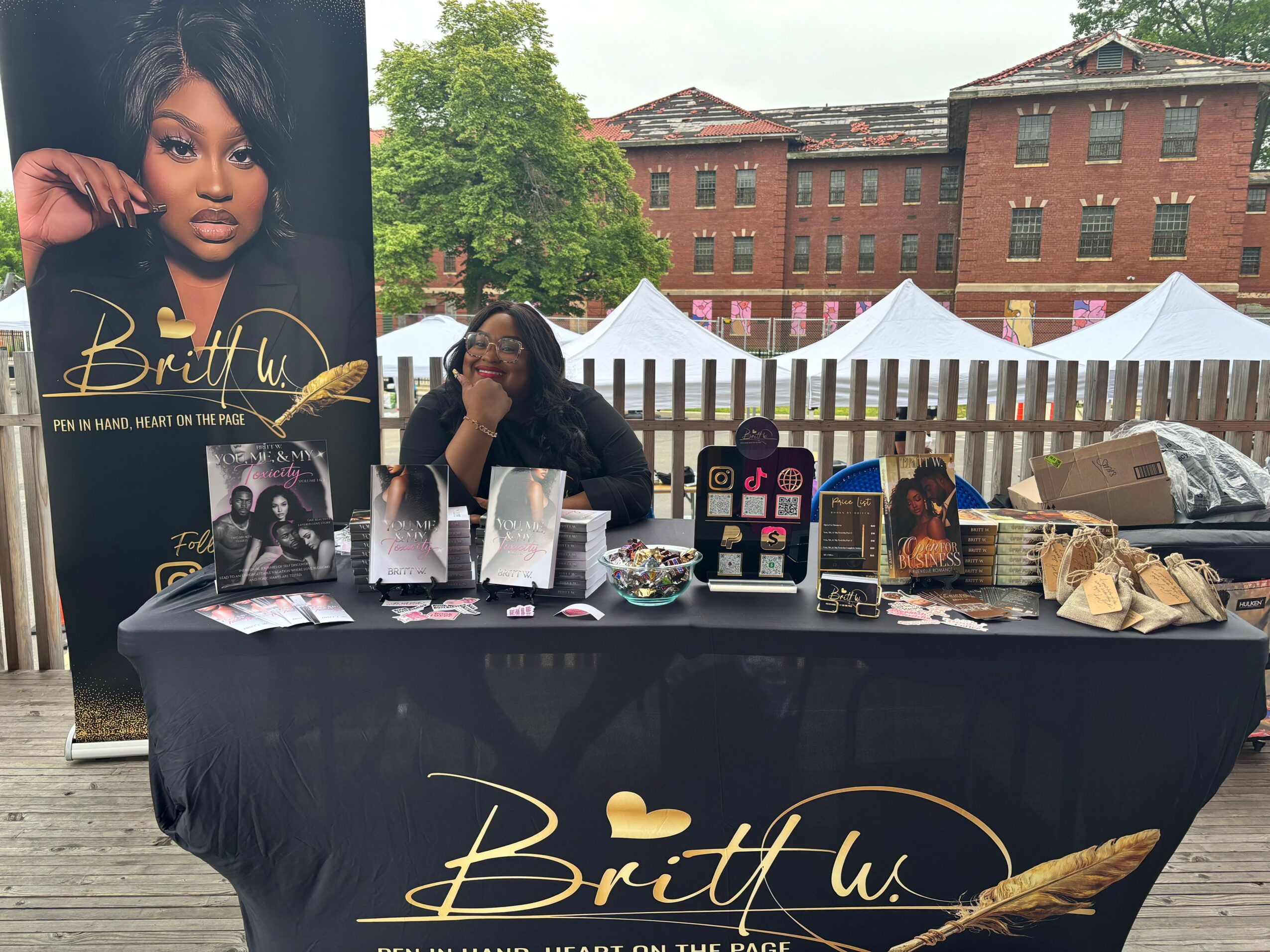 Beauty booth with promotional poster of a woman, table with products, and outdoor tents in background.