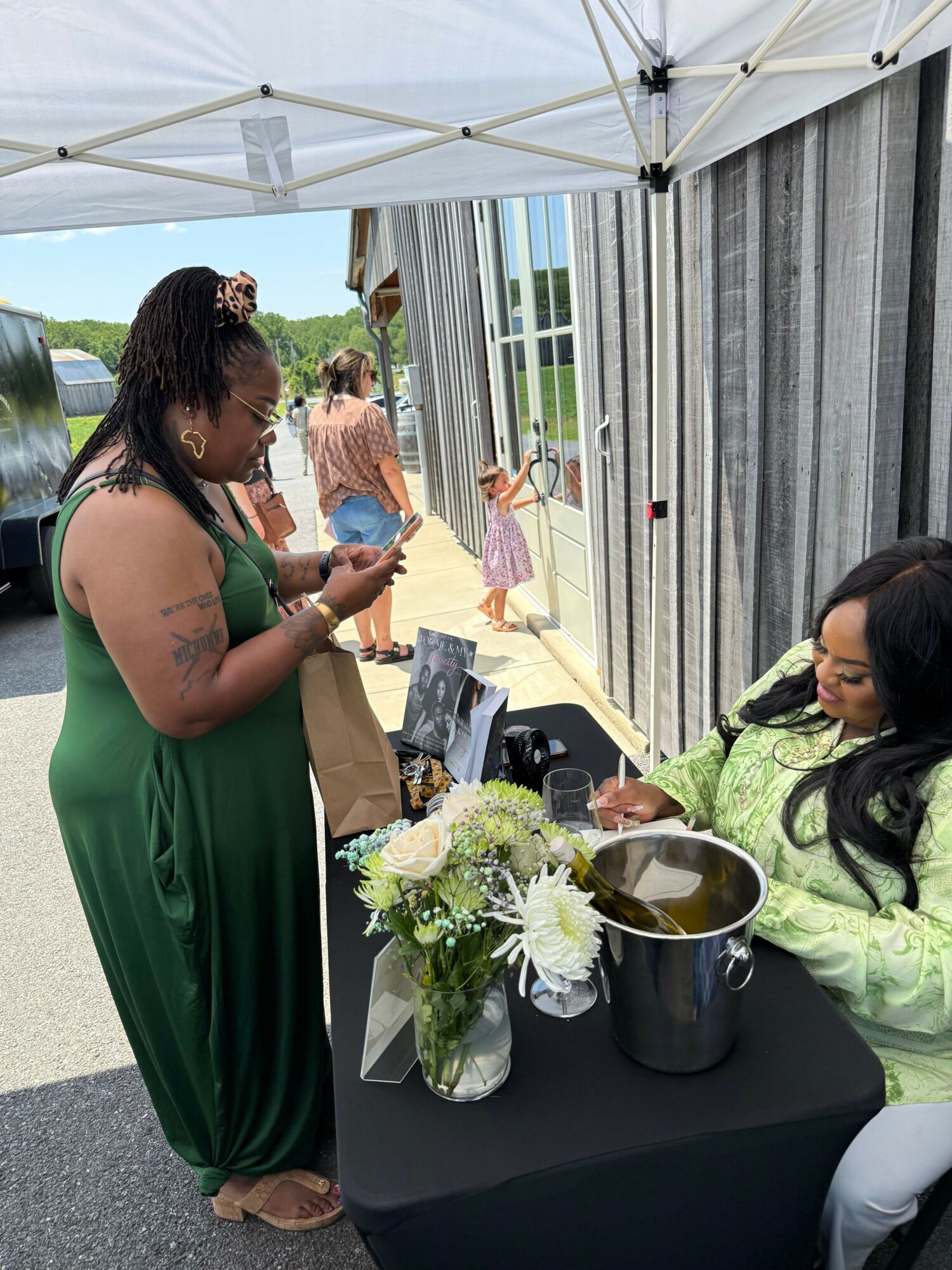 Two women at an outdoor event, one pouring a drink into a glass, the other looking on, with a table and flowers.