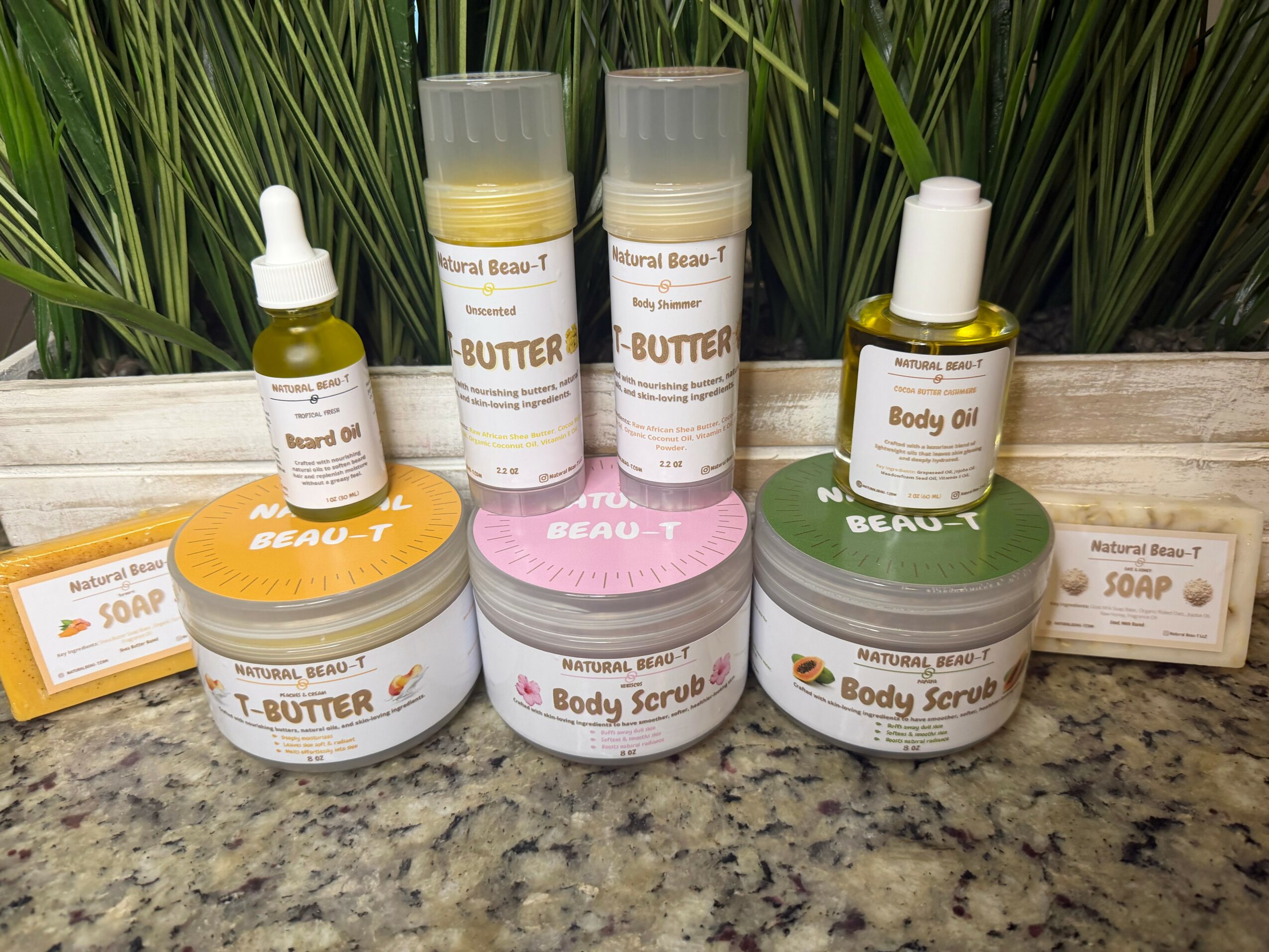 Skincare products including body butter, body scrub, soap, and body oil arranged on a wooden surface with green plants in background.