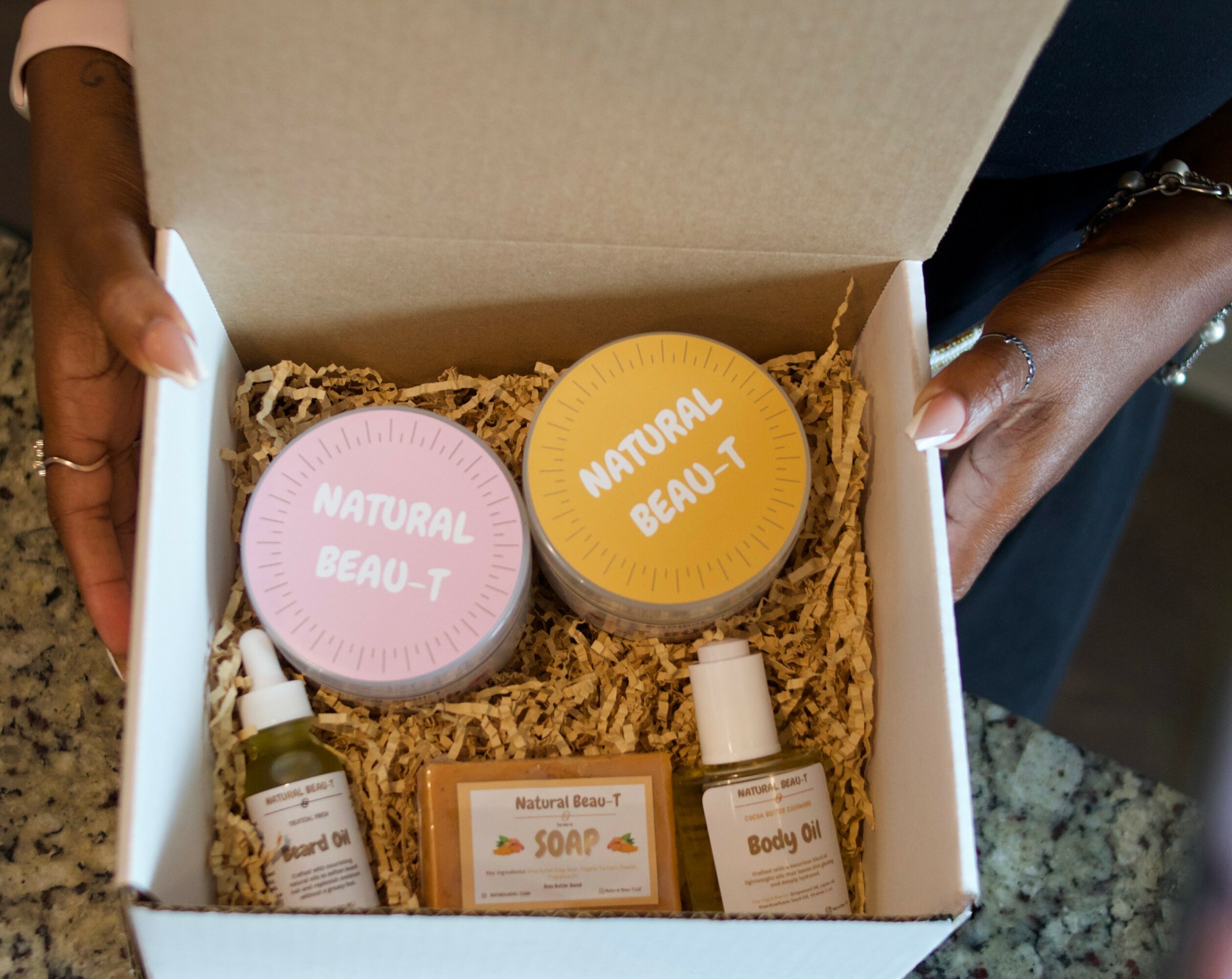 Open box with natural beauty products, including two jars, soap, and body oil, on shredded paper filler.