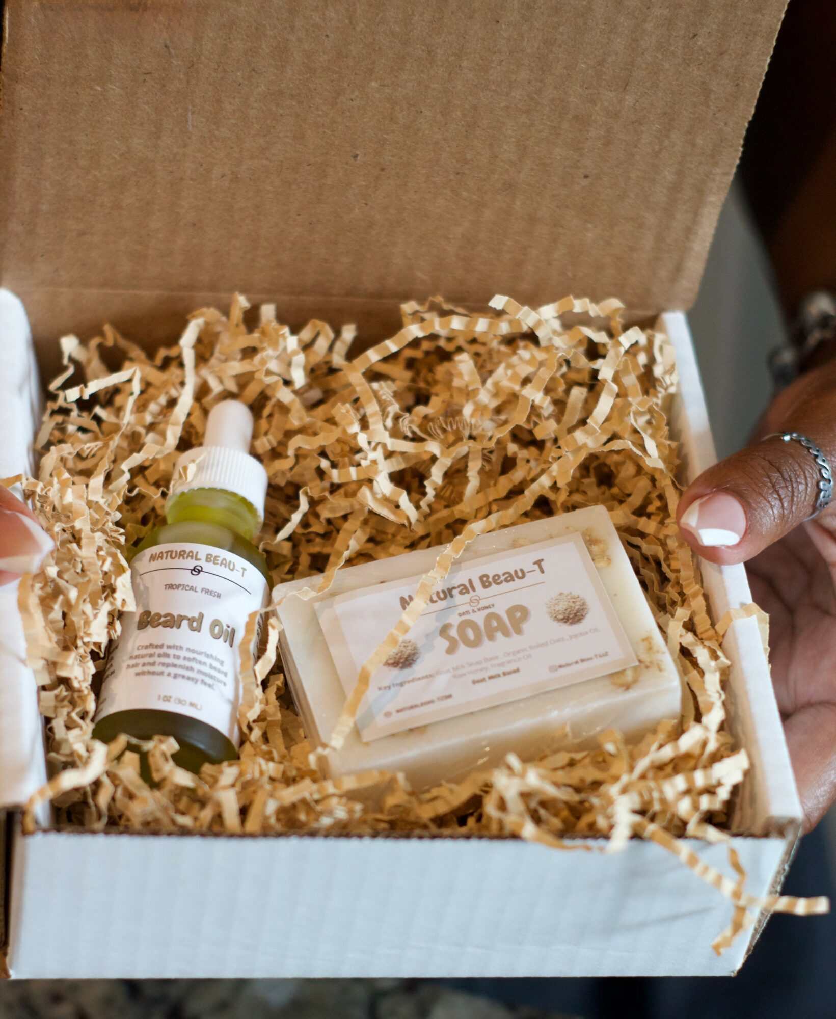 Open box with soap and a small bottle, filled with paper shred filler, held by two hands with rings.