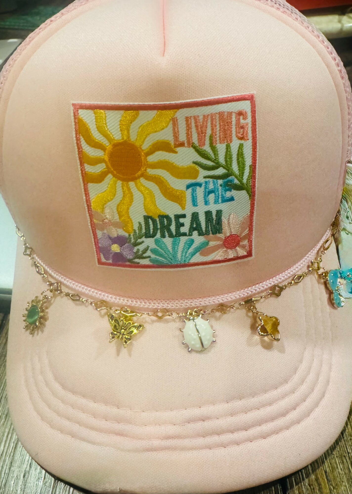 Pink cap with embroidered sun, plants, and text reading 'Living the Dream,' and a chain of charms hanging from the brim.