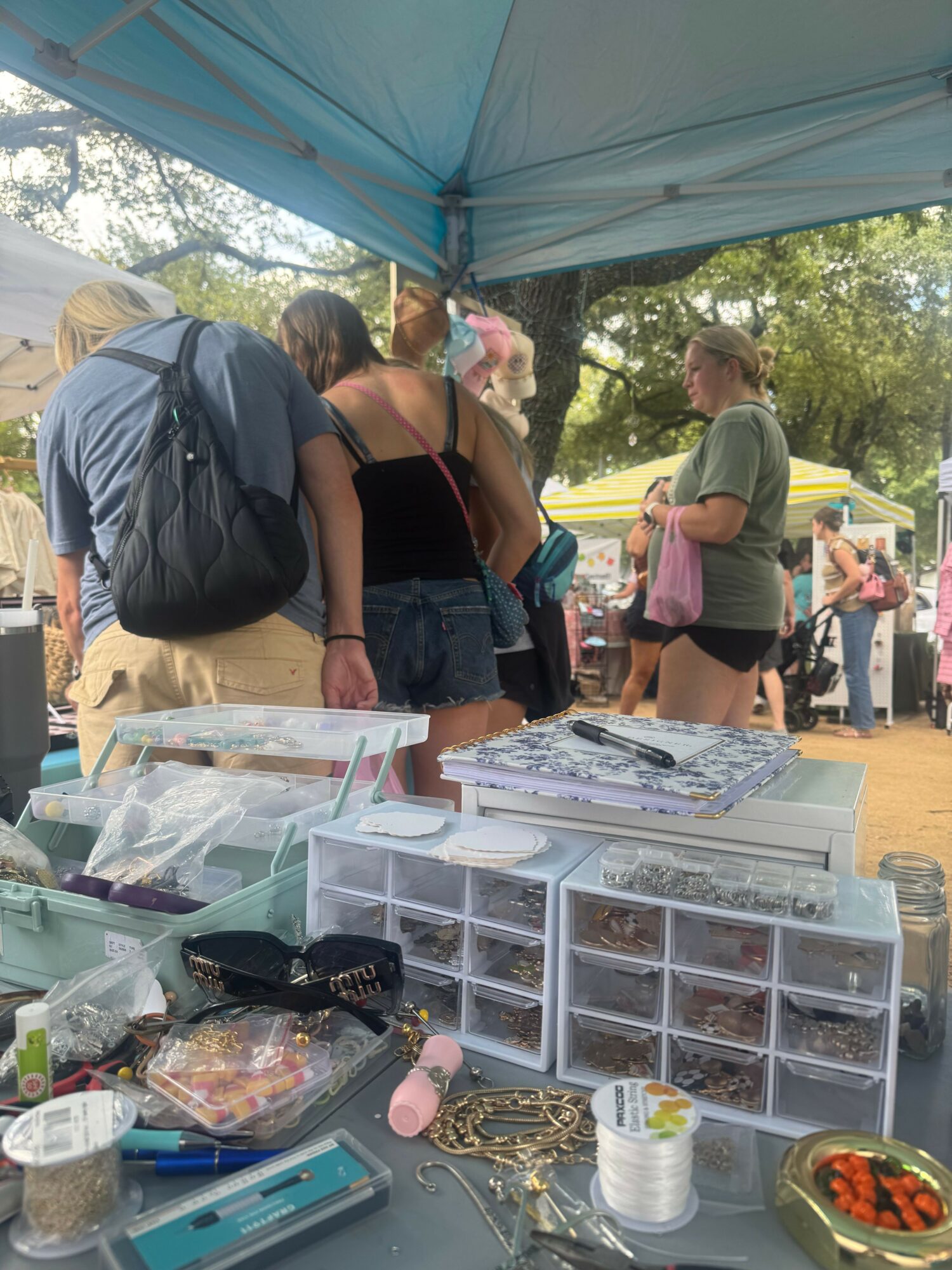 People browsing items at an outdoor market stall with various containers and supplies on a table.
