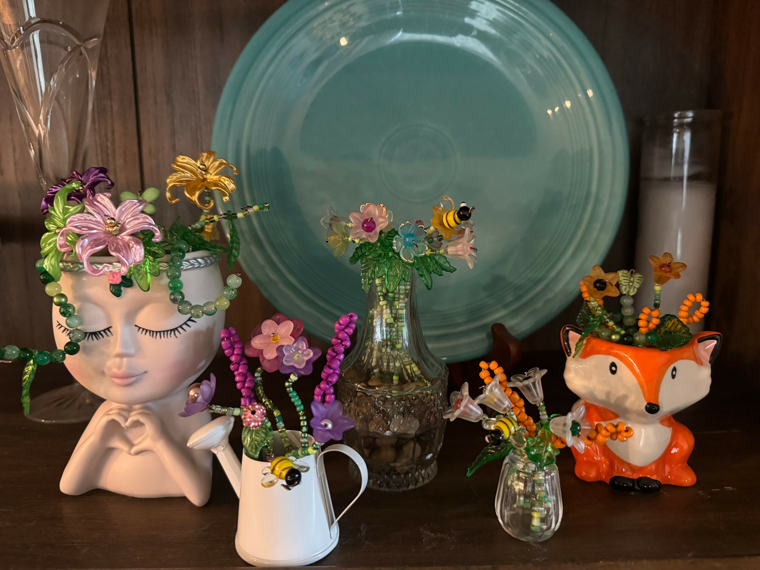 Decorative display with a mannequin head, flowers, bees, a large plate, and a fox figurine on a wooden surface.