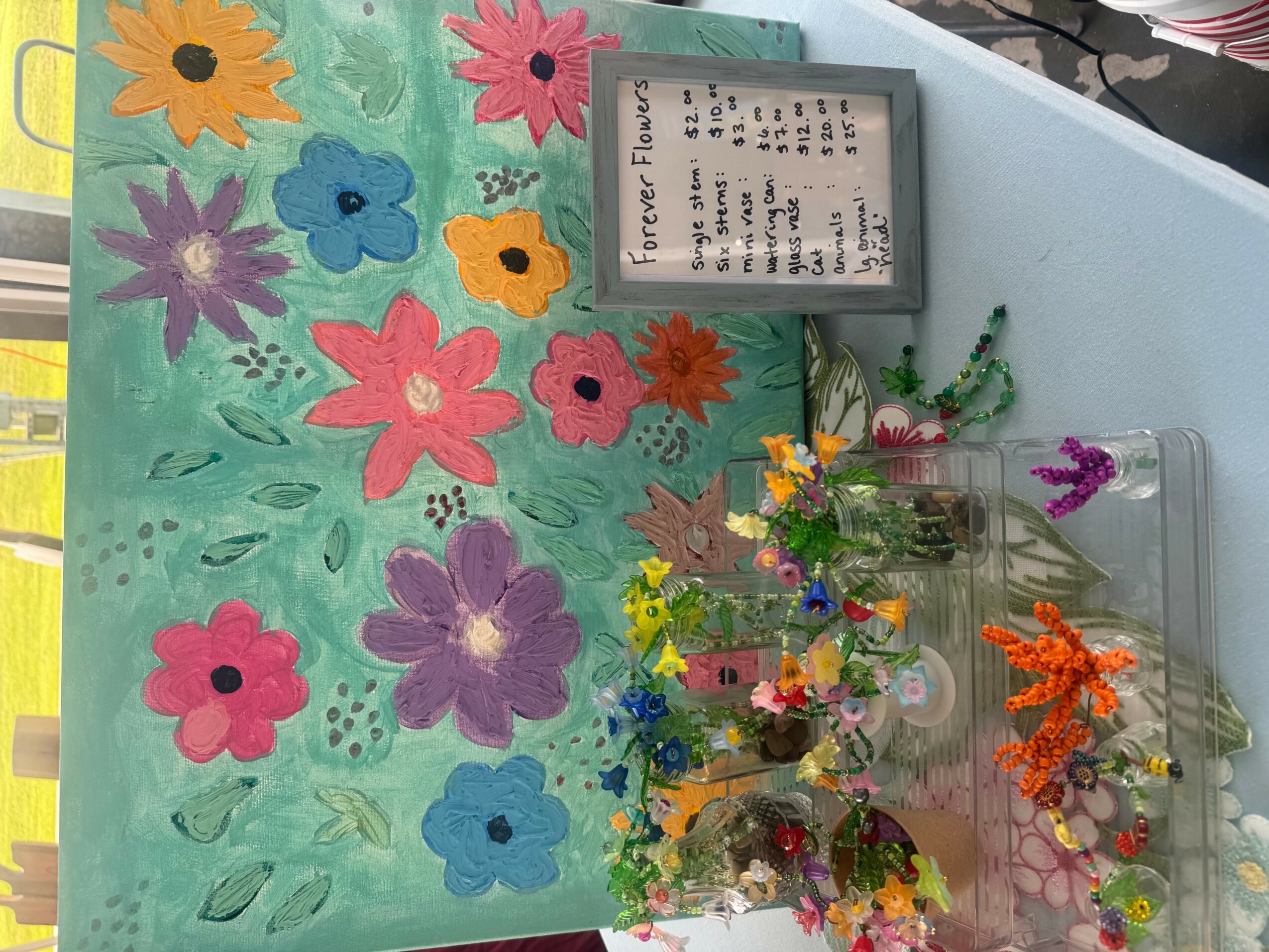 Colorful painted flowers on a green background with small flower arrangements in glass containers, and a framed sign, all on a table.