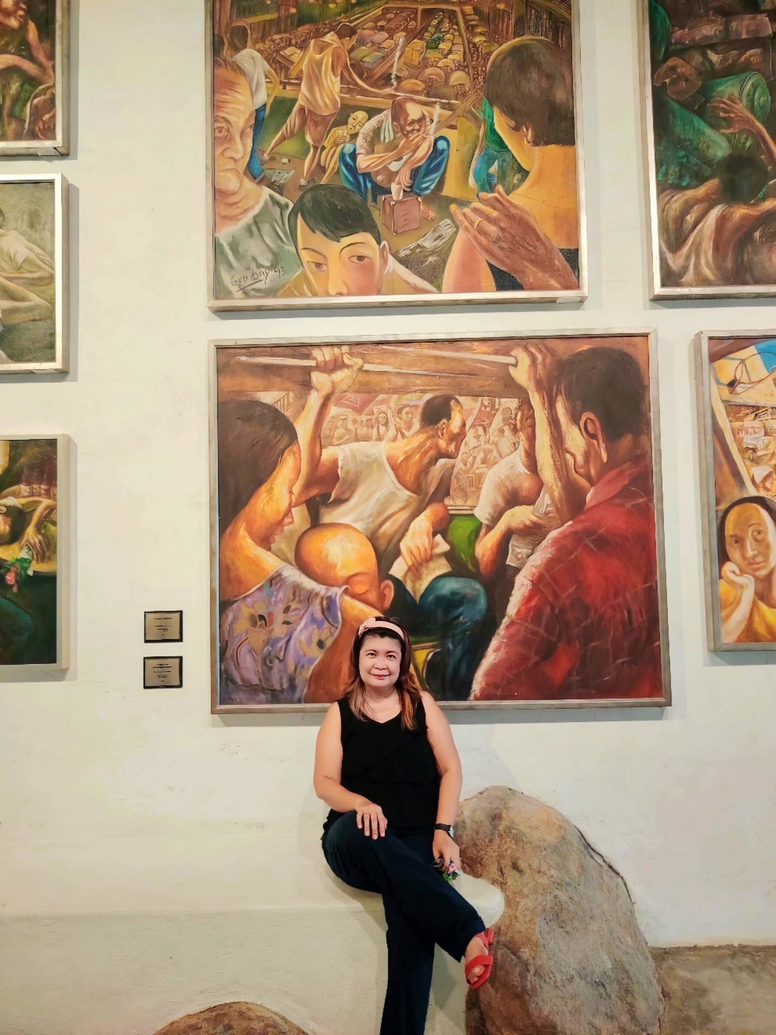 Young woman with black hair and glasses sitting on a large rock in front of colorful paintings on a gallery wall.