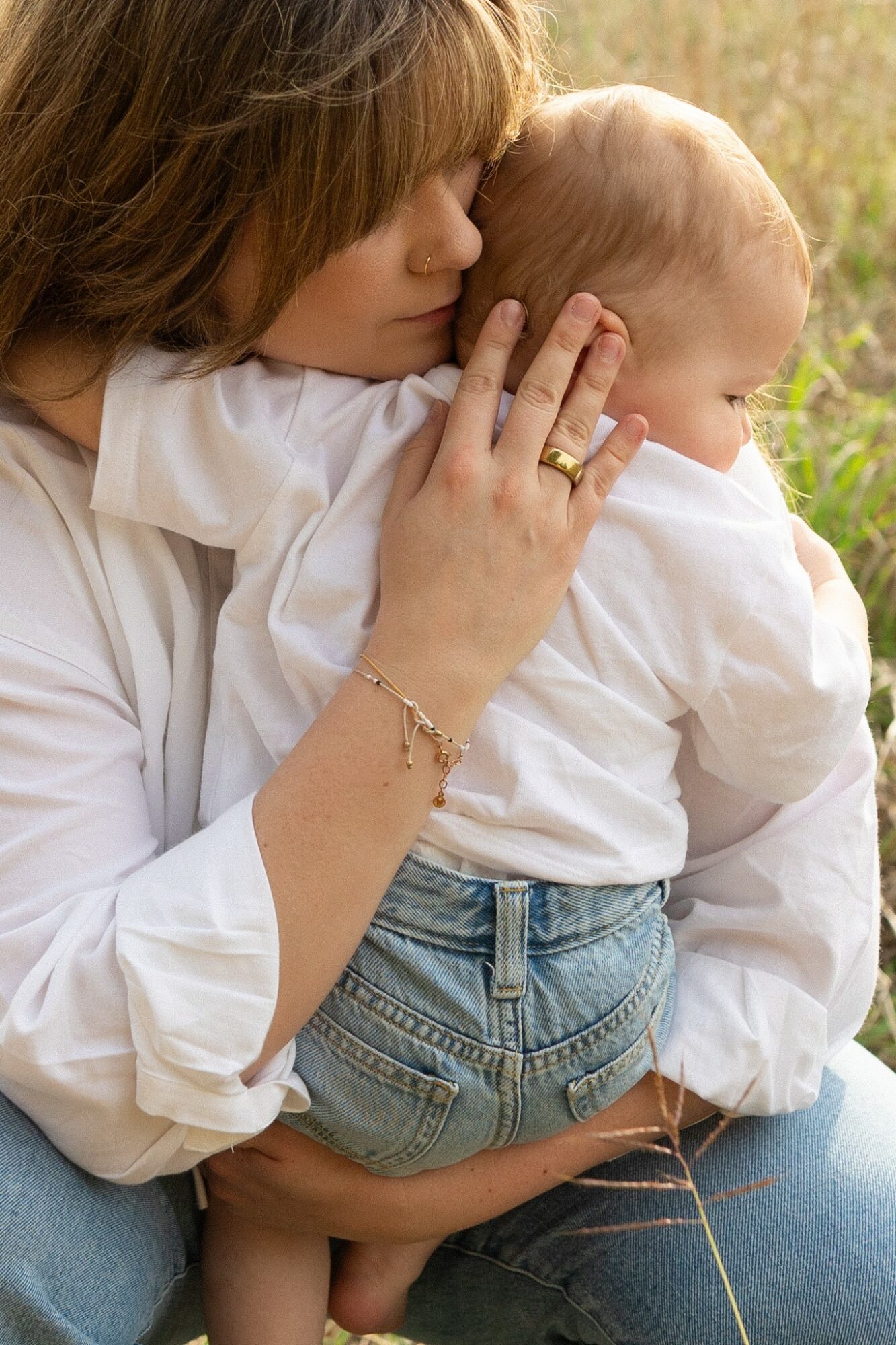 Woman holding a baby outdoors, close-up view, woman wearing a white shirt, baby in denim shorts, natural background.