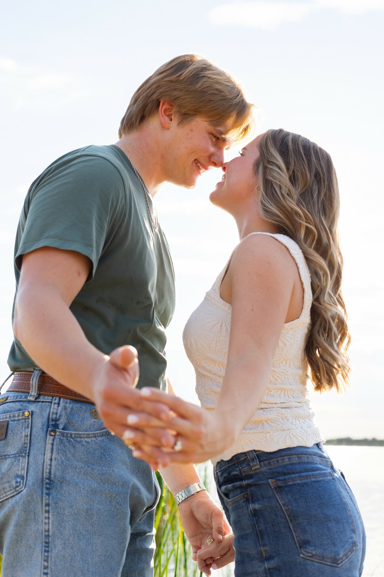 A couple holding hands, smiling, with their foreheads touching outdoors against a bright sky.