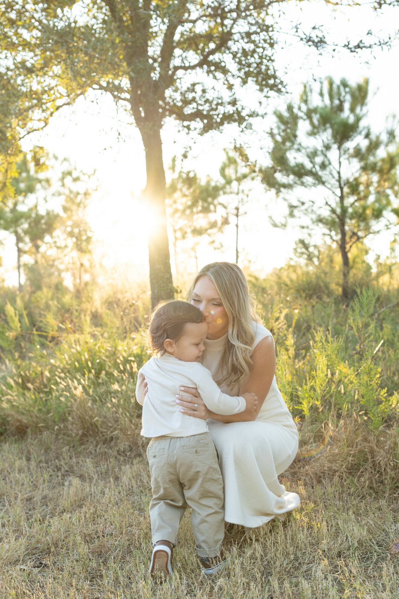Woman kneeling with child outdoors in sunlight, trees in background, grassy area, warm lighting, peaceful scene.