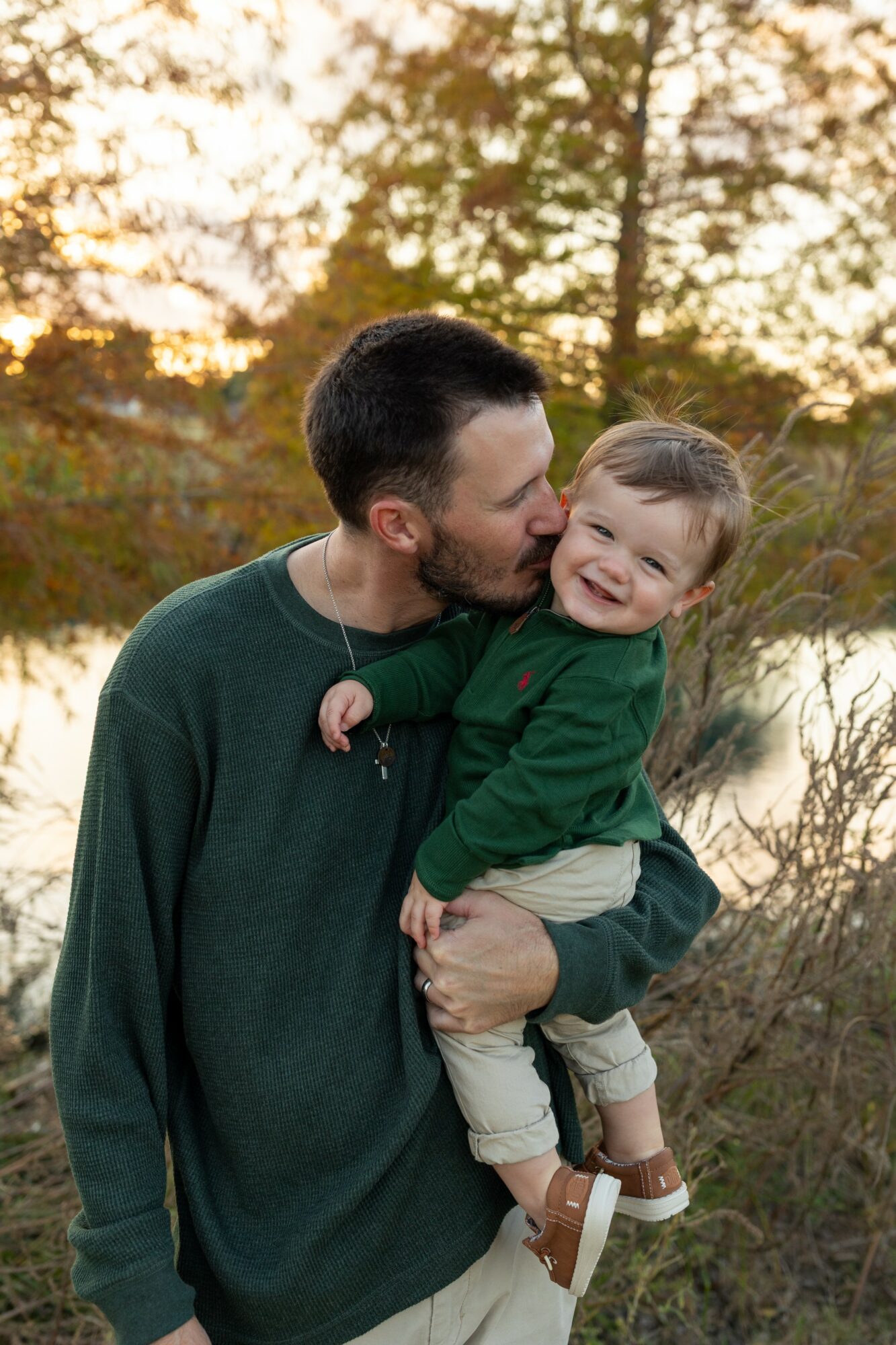 Man holding smiling child outdoors during sunset, trees and water in background.
