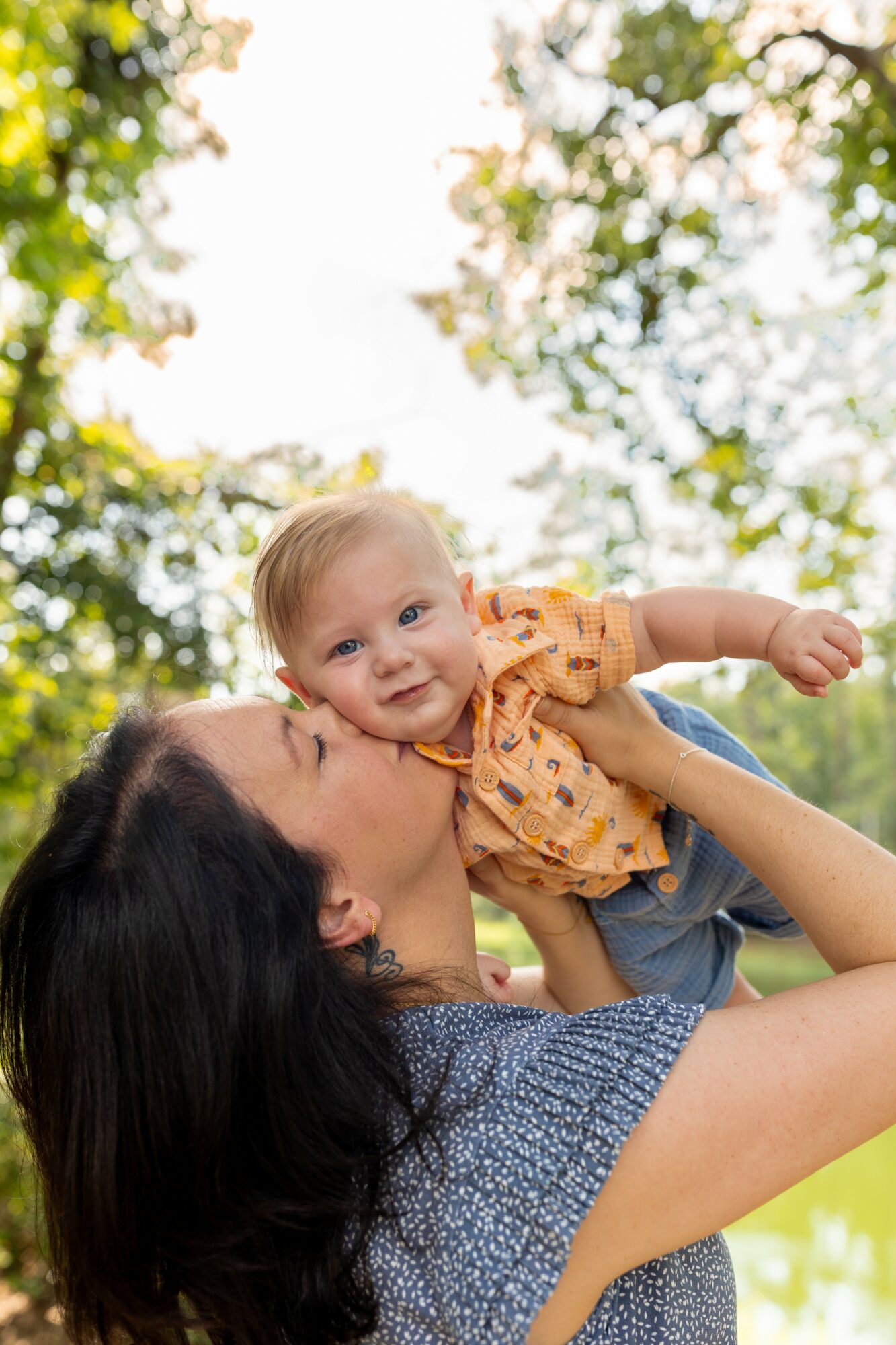 Woman holding a smiling baby outdoors with trees and sunlight in background.
