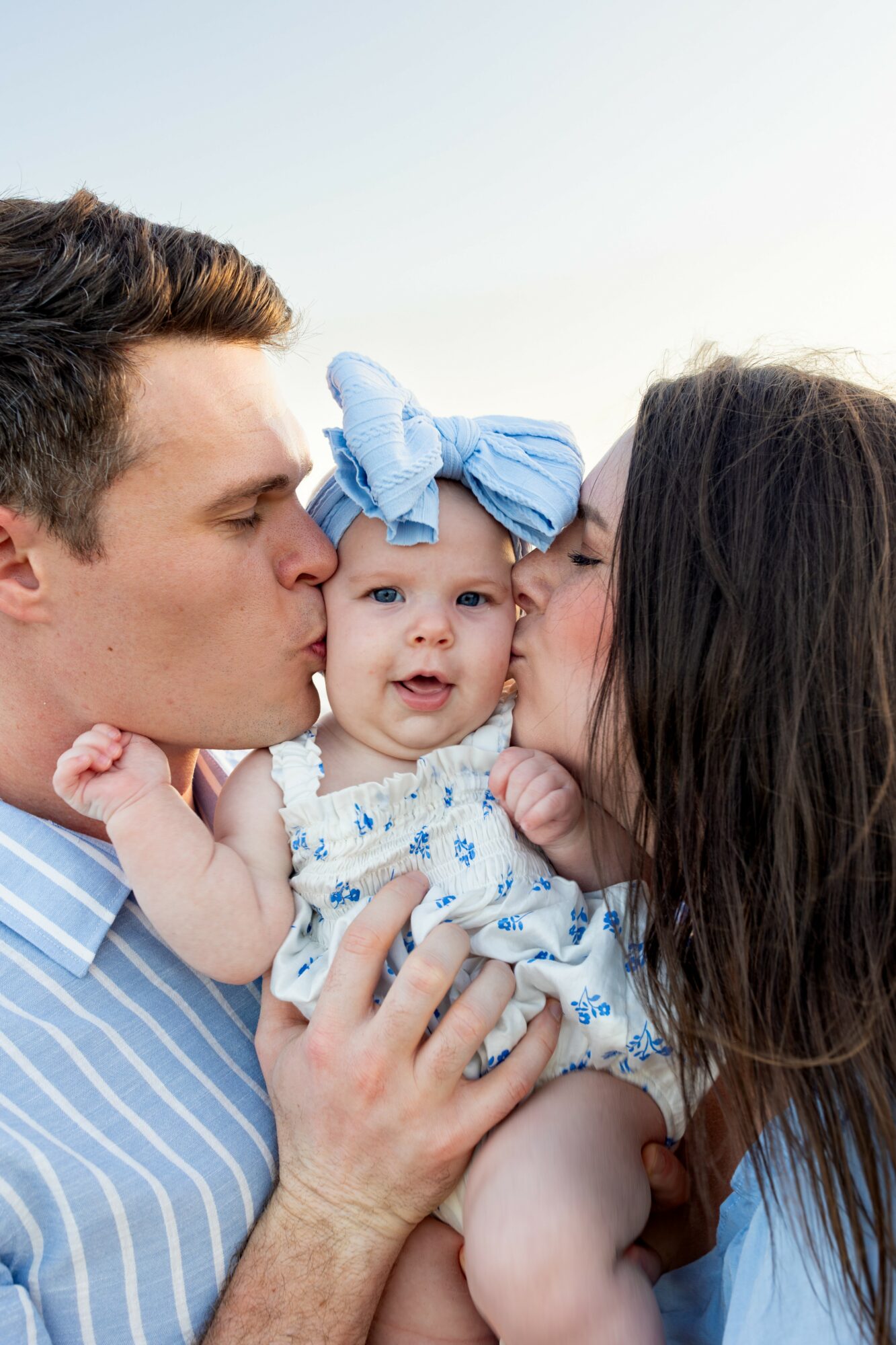 Man and woman kiss a baby girl with a large blue bow on her head, outdoors against a light sky.