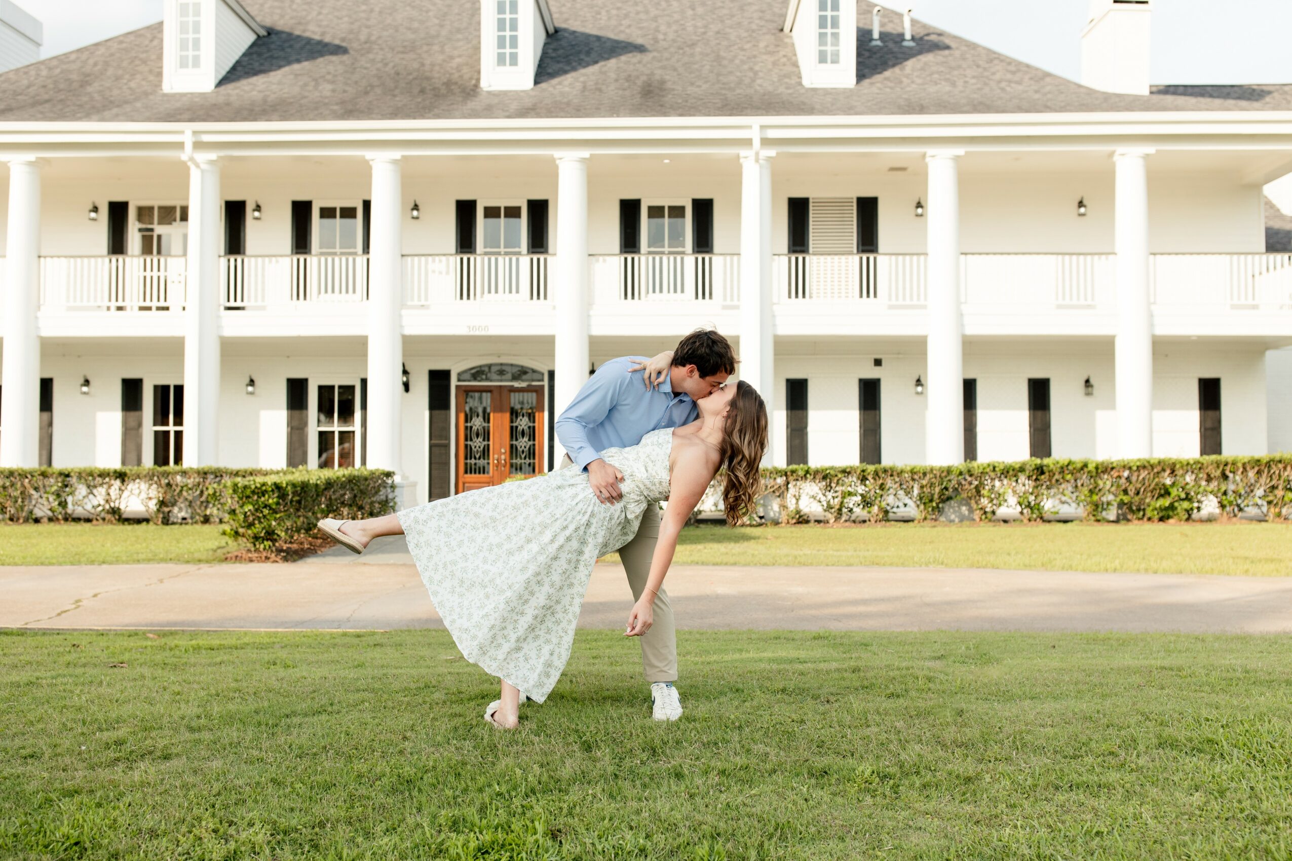 Couple dancing on grass in front of a large white building with balconies, woman in a light dress, man in a blue shirt.