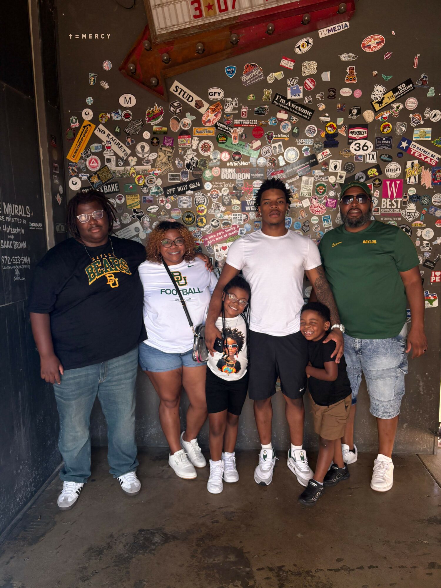 Group of five people standing in front of a wall covered with stickers and patches, some holding a microphone.