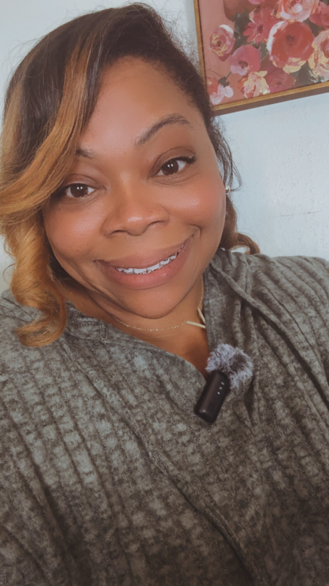 Smiling woman with shoulder-length hair wearing a gray sweater, indoors with a floral picture in the background.