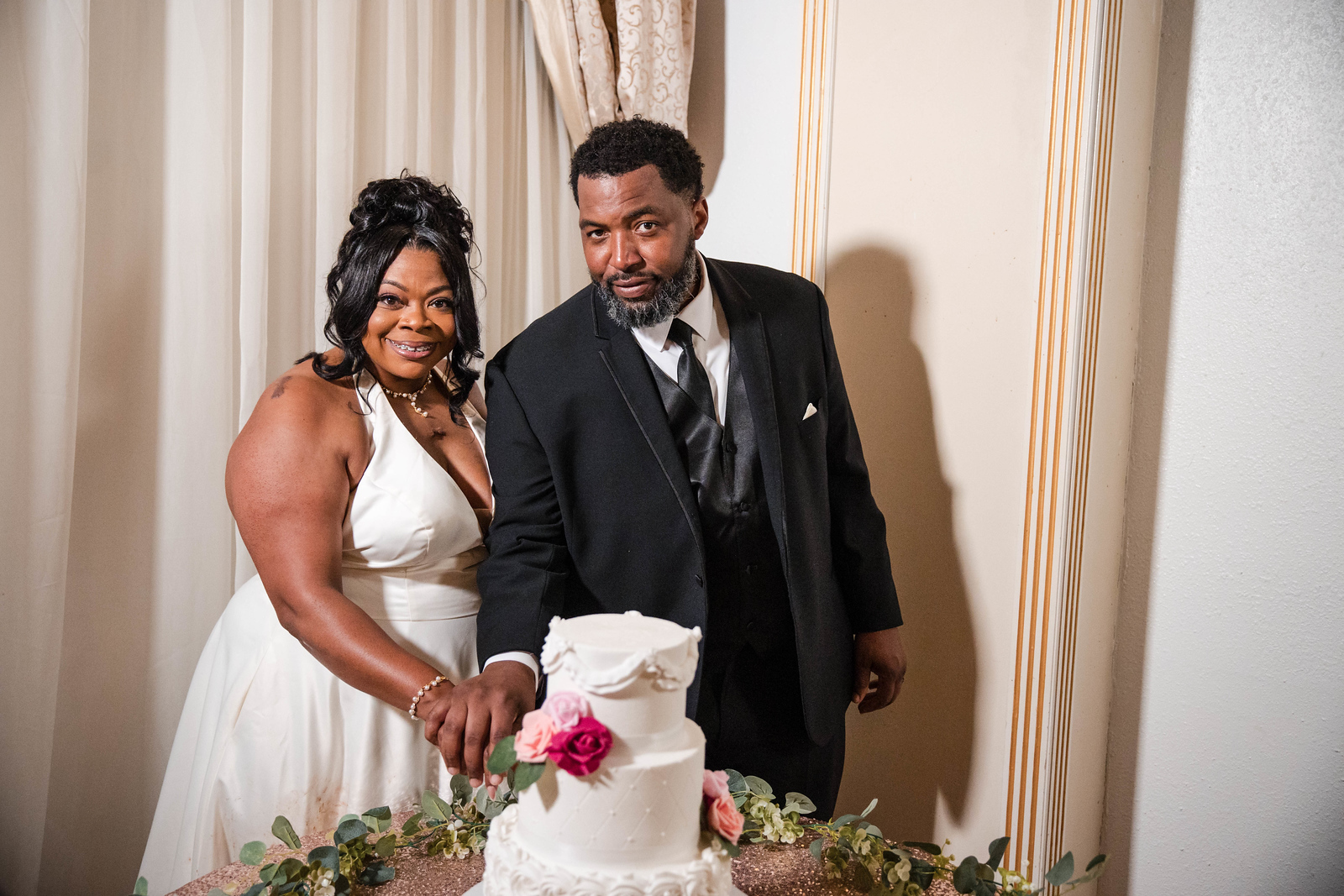 A smiling woman in a white dress and a man in a black suit stand together at a celebration with a cake and flowers.