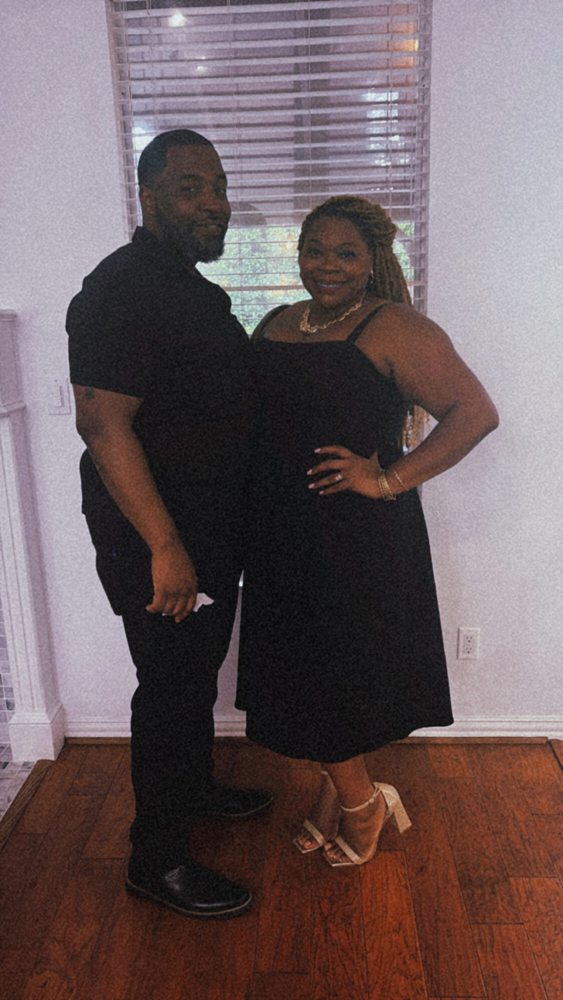 A man and woman stand together indoors, smiling, with a window and blinds behind them. The woman wears a black dress and heels.