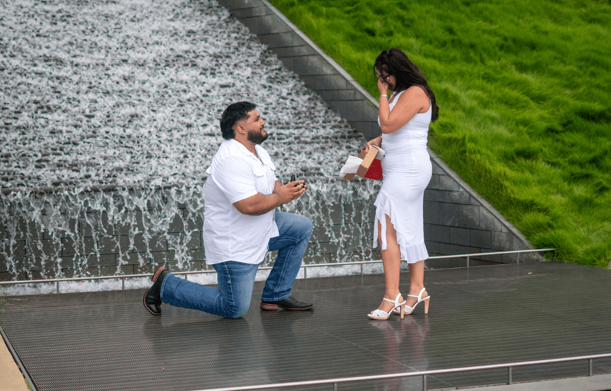 Man kneeling on one knee proposing to woman holding a gift, near a waterfall and green grass