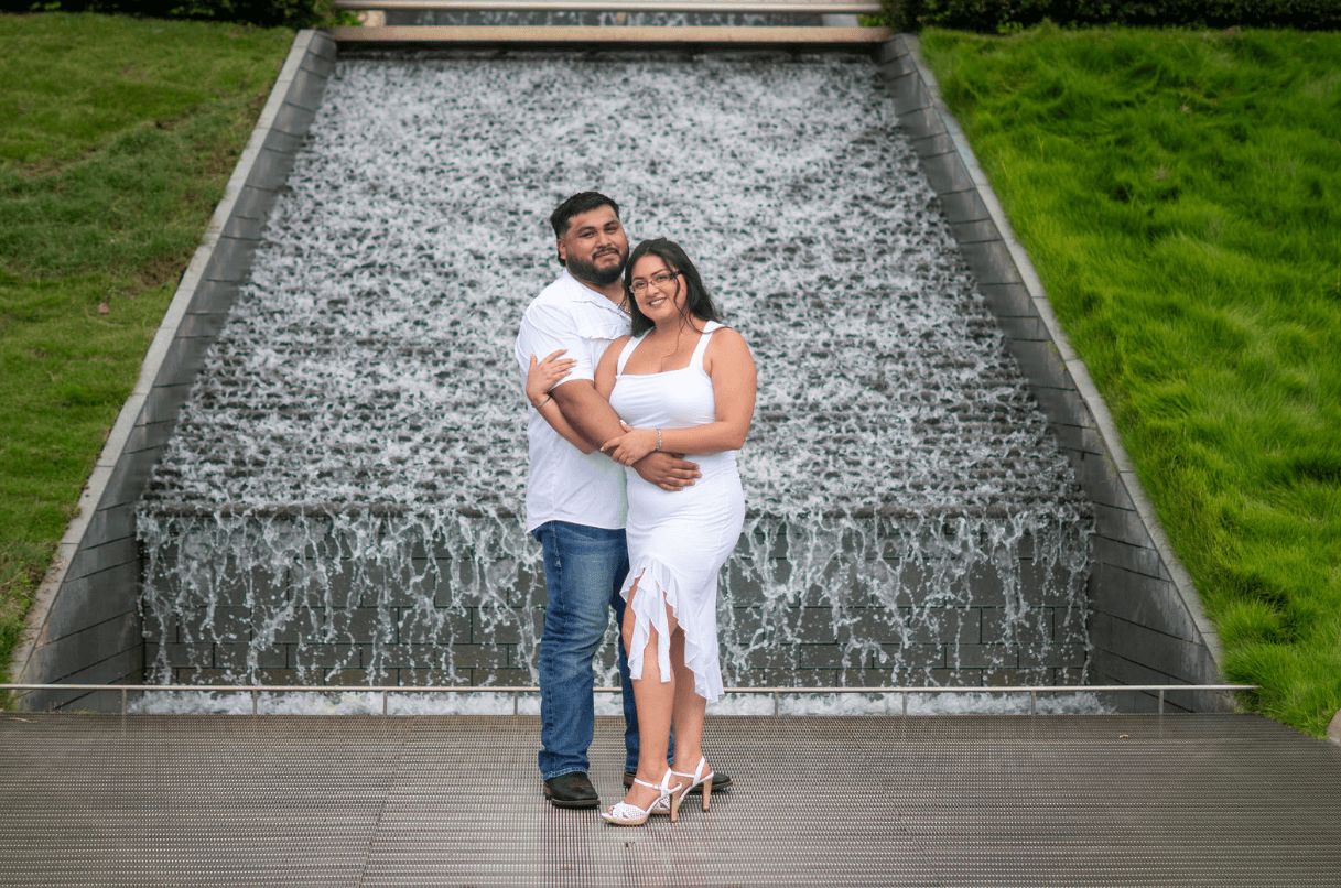 A couple stands in front of a cascading water feature with green grass on both sides, smiling and embracing.