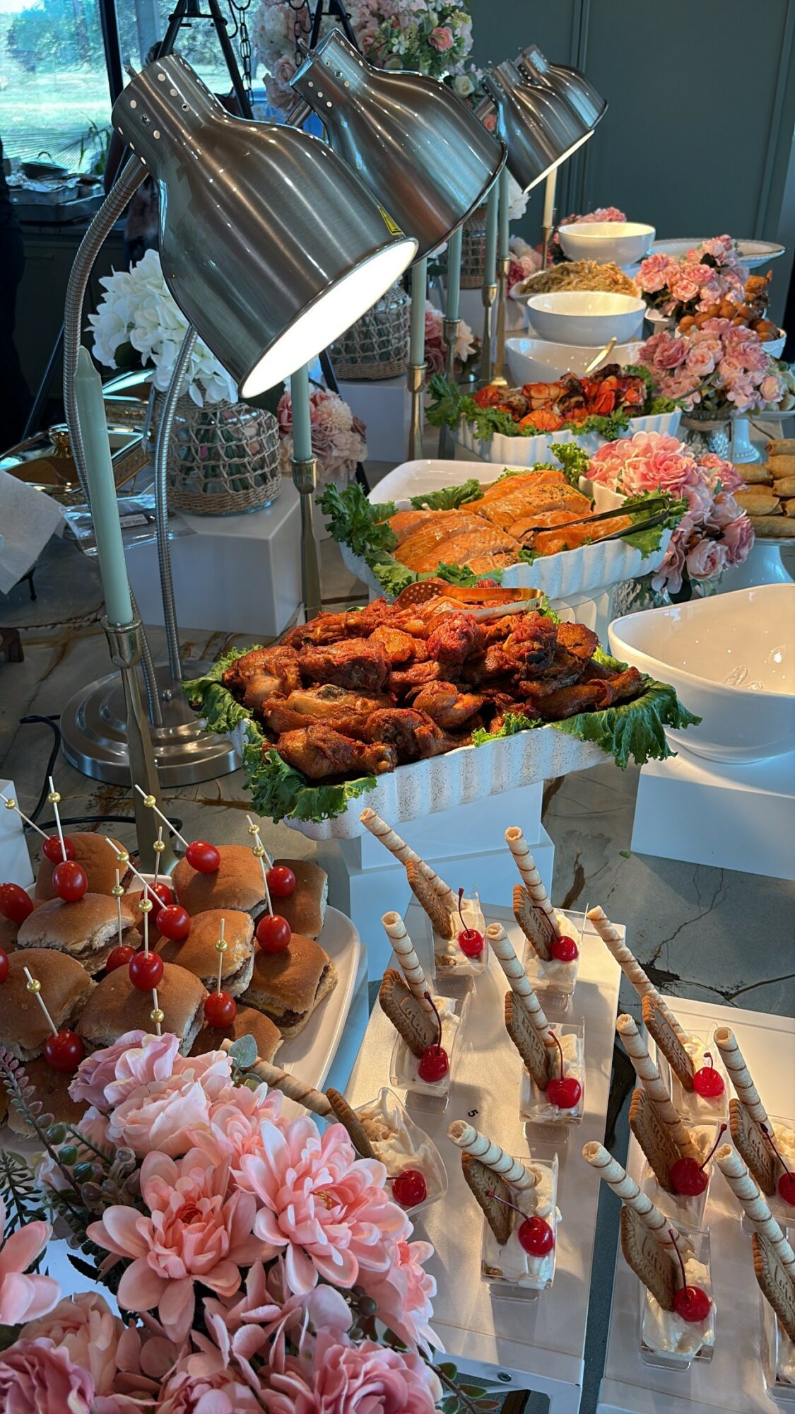 Assorted desserts including cupcakes, cake pops, and pastries displayed on a table with flowers and lamps.