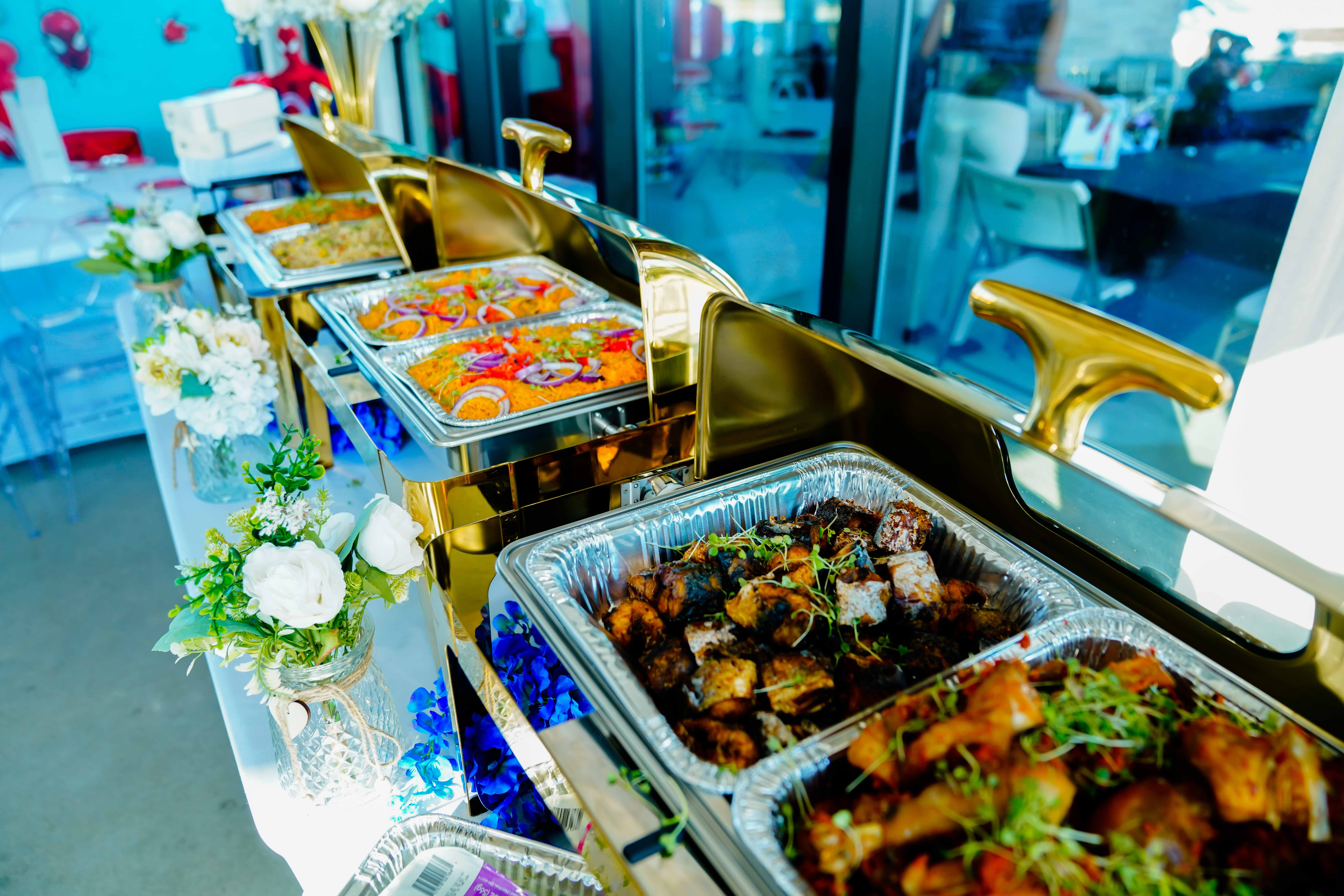Buffet table with trays of cooked food, surrounded by white flowers and gold serving utensils, in a bright room.