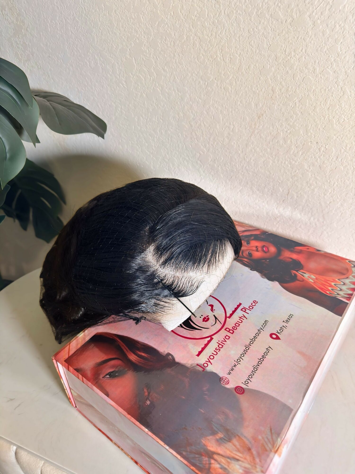 A black and white wig rests on a magazine with a woman's face on the cover, placed on a white surface.