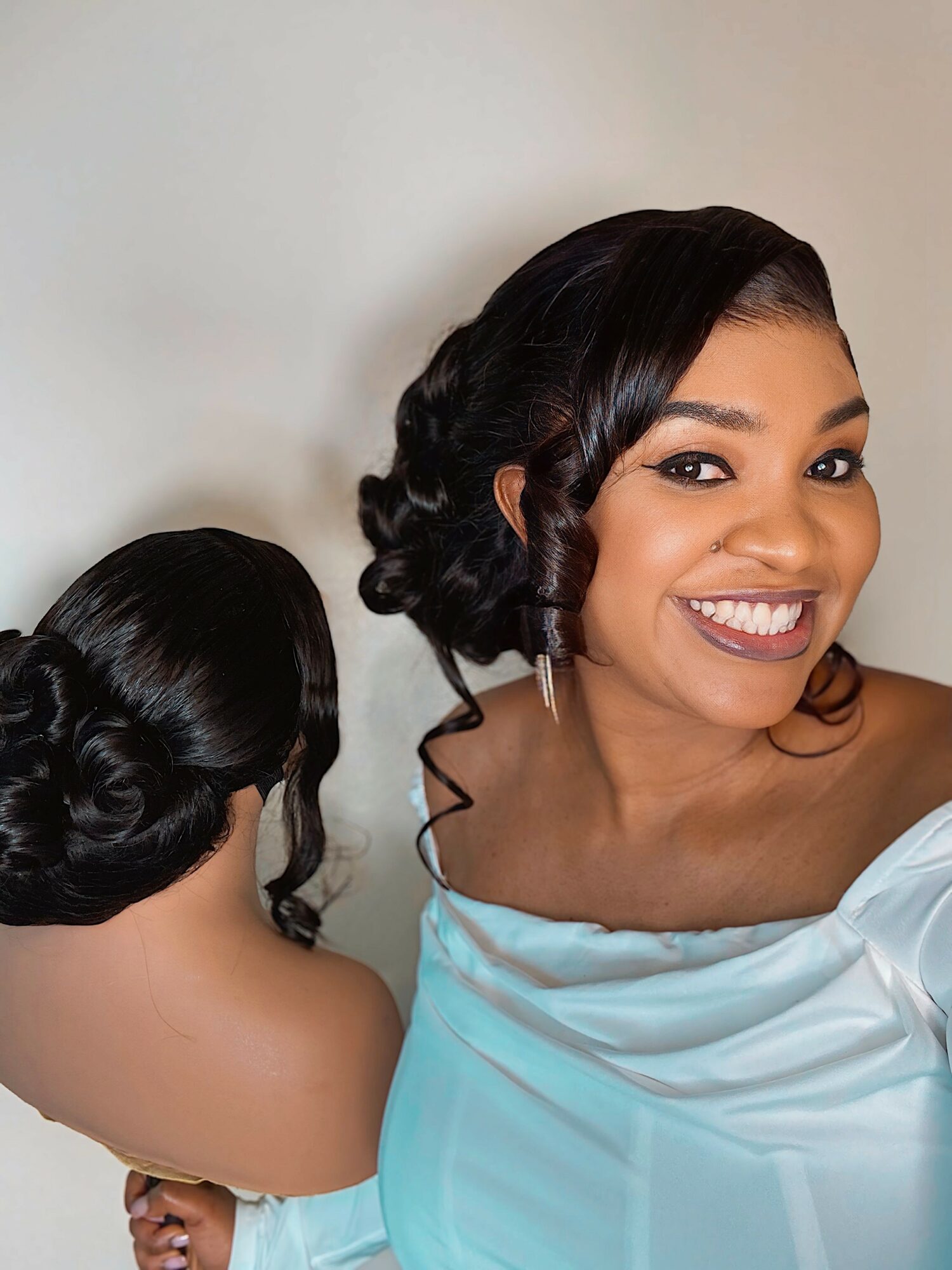 Smiling woman with styled hair wearing a white off-shoulder top, facing camera, with a neutral background.