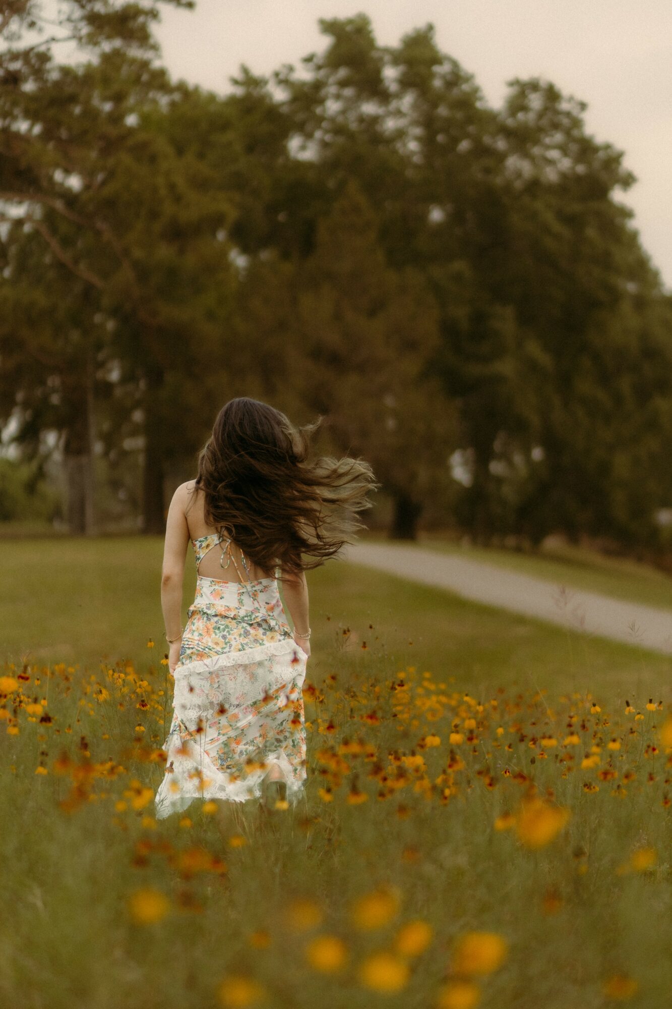 Girl with long hair walking through a field of yellow flowers, trees in the background, wind blowing her hair.