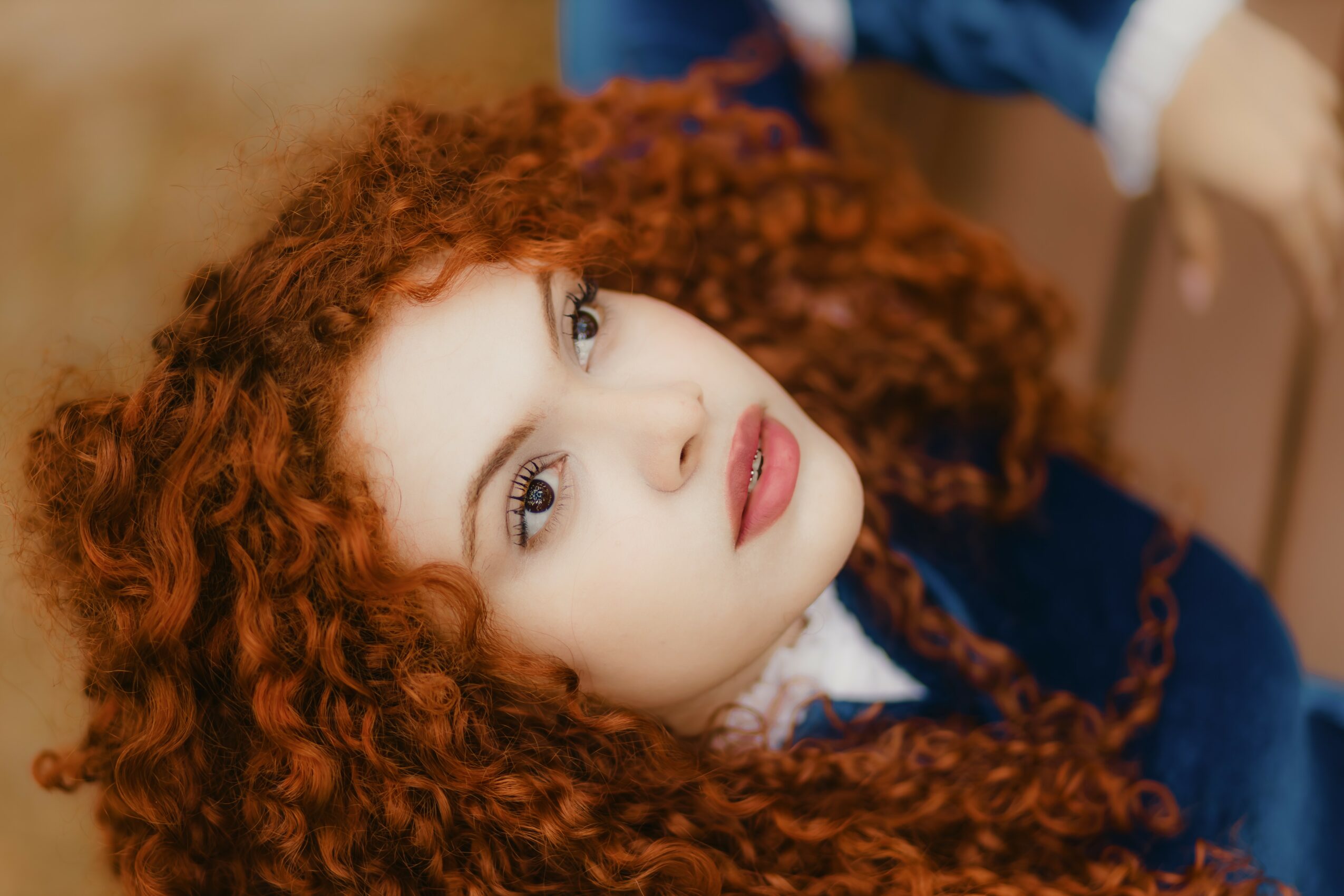 Young woman with curly red hair and light makeup, looking directly at the camera, wearing a dark blue outfit.