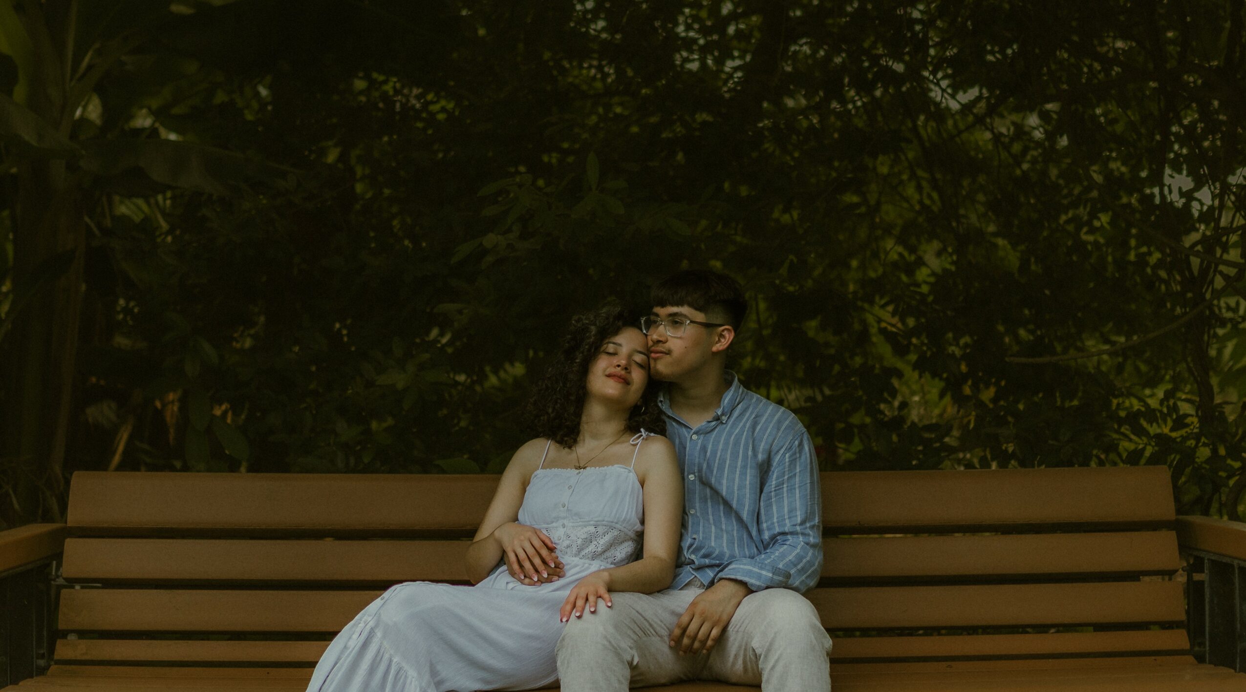 A couple sitting on a park bench, with trees in the background, the woman in a white dress and the man in a blue shirt.