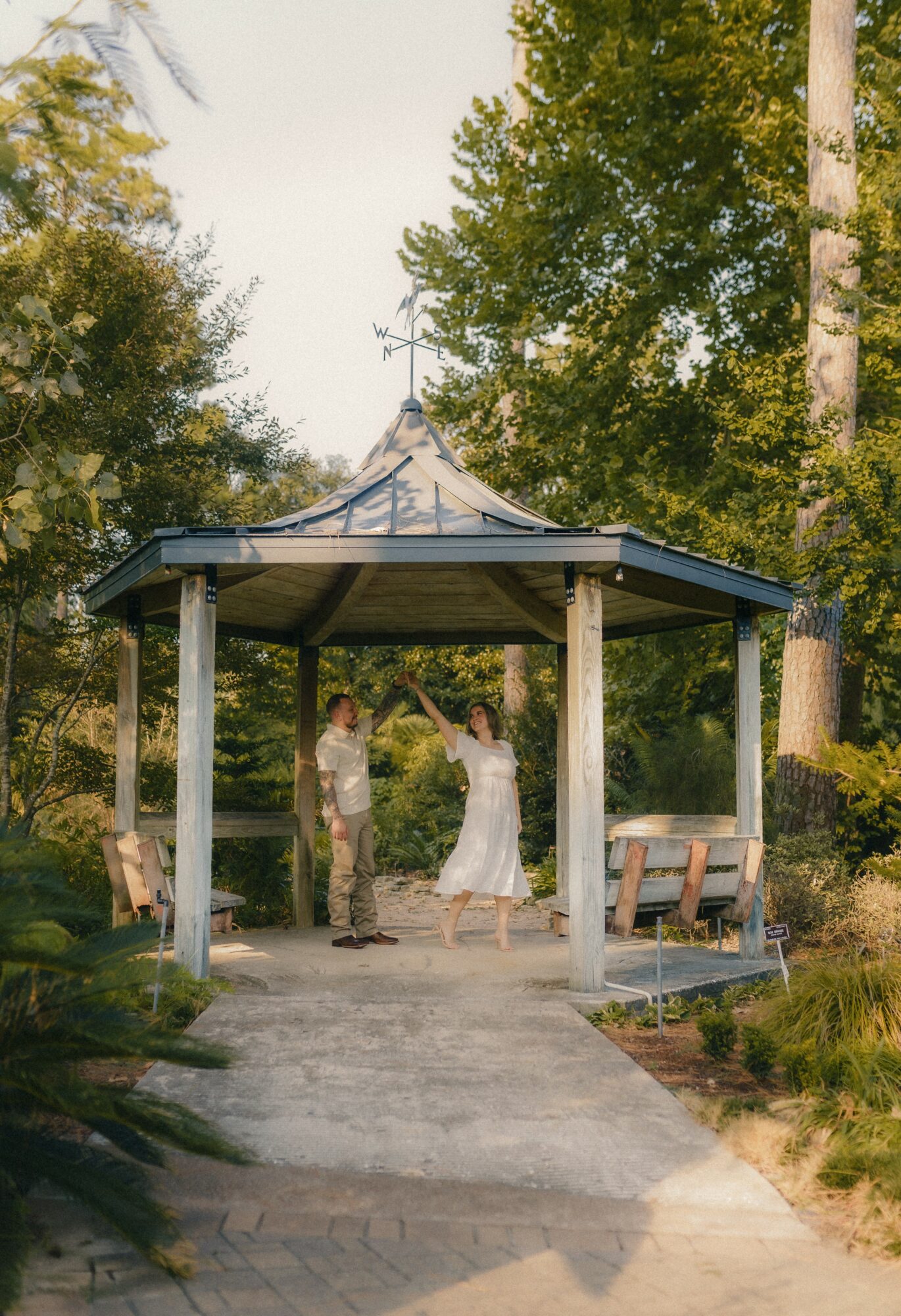 Two people stand under a gazebo in a park, surrounded by trees and benches, with sunlight filtering through leaves.