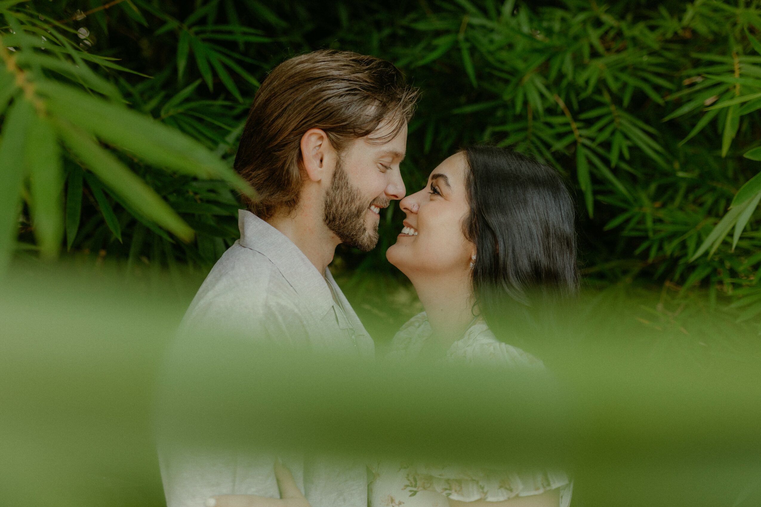 A couple faces close together, smiling, surrounded by green foliage and large leaves.