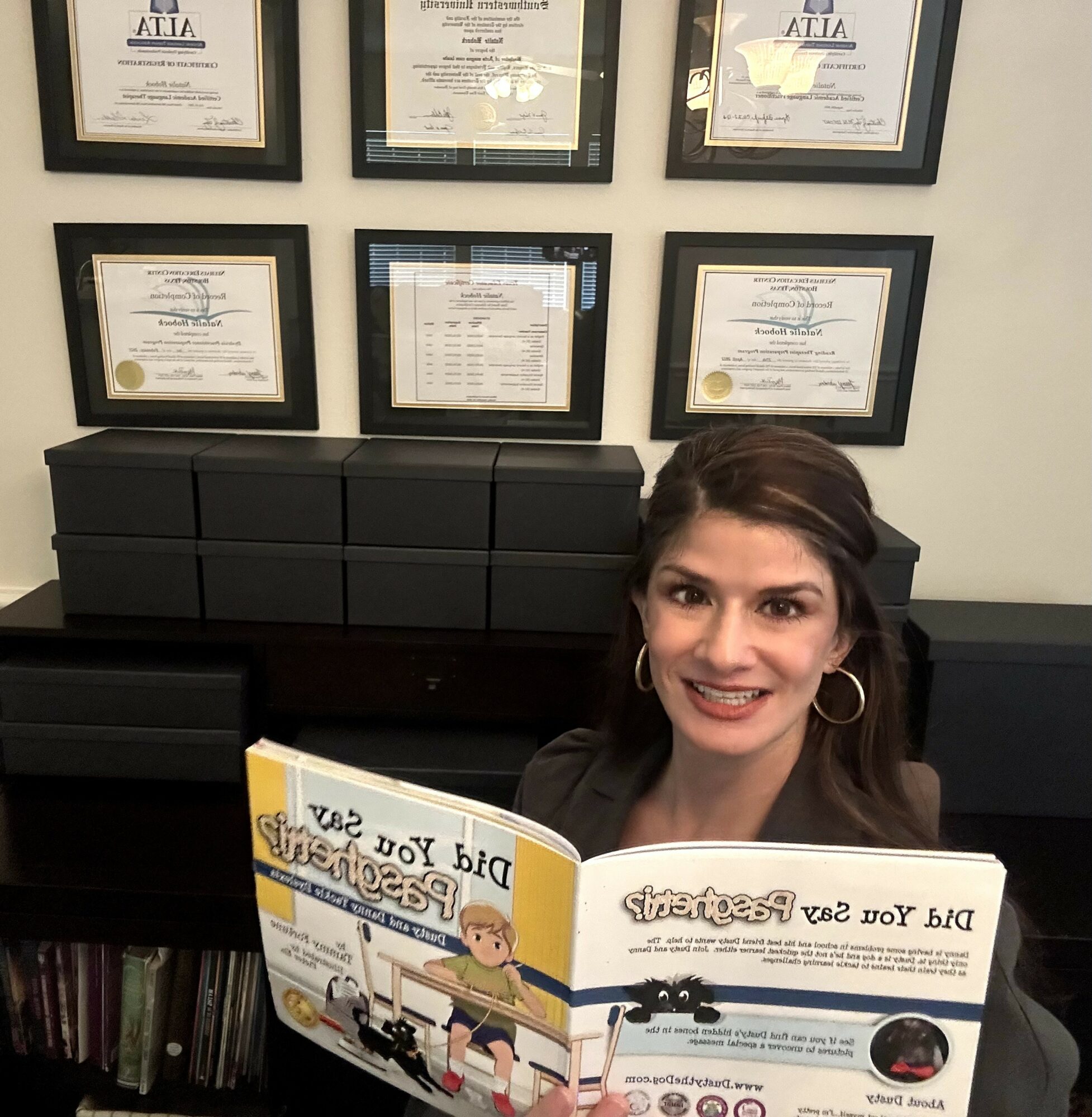 Young woman with dark hair and hoop earrings reading a children's book, smiling, in a room with framed certificates on the wall.
