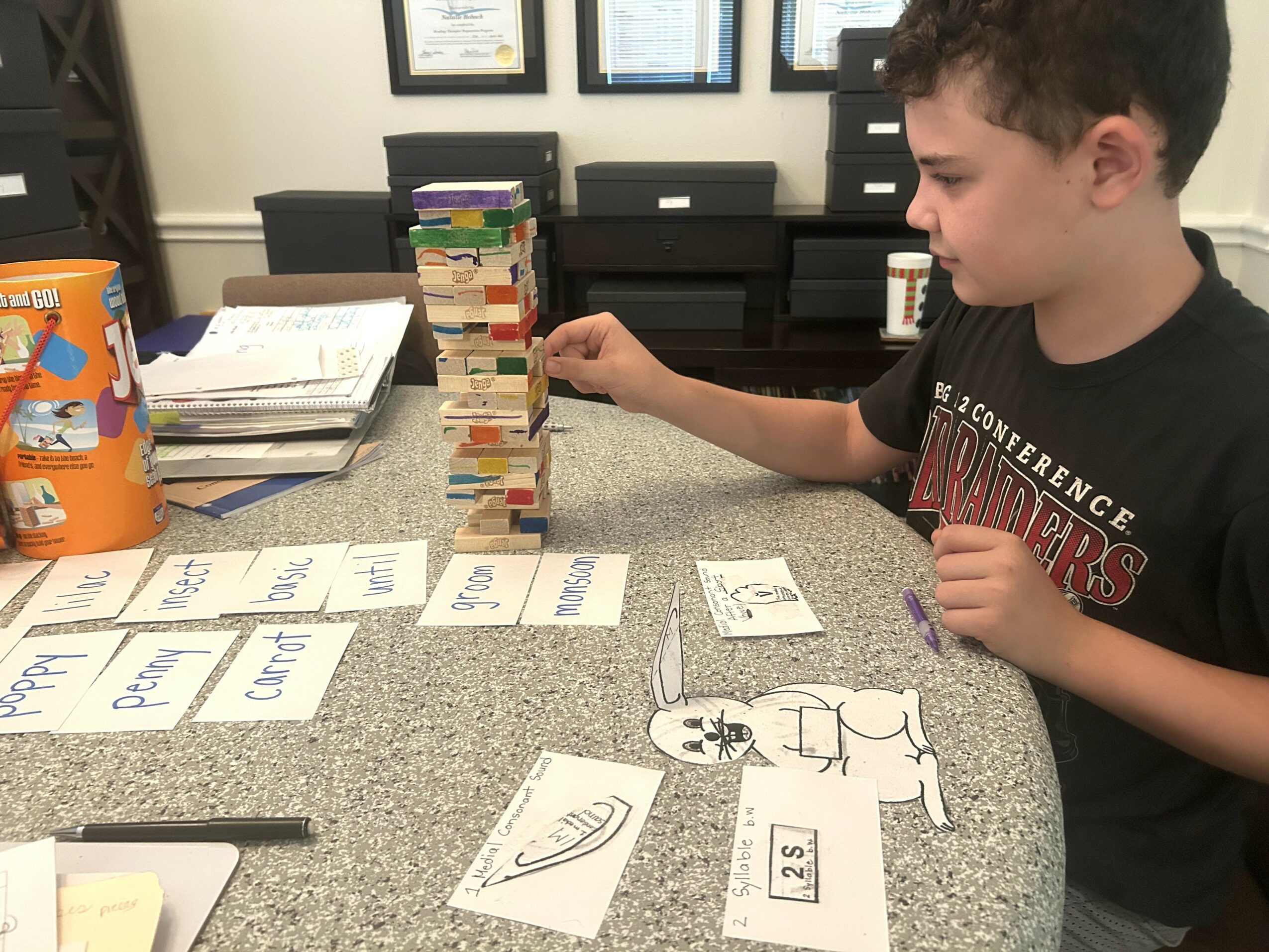 A boy playing a card game with a Jenga tower on a table, papers, and a cup in the background.