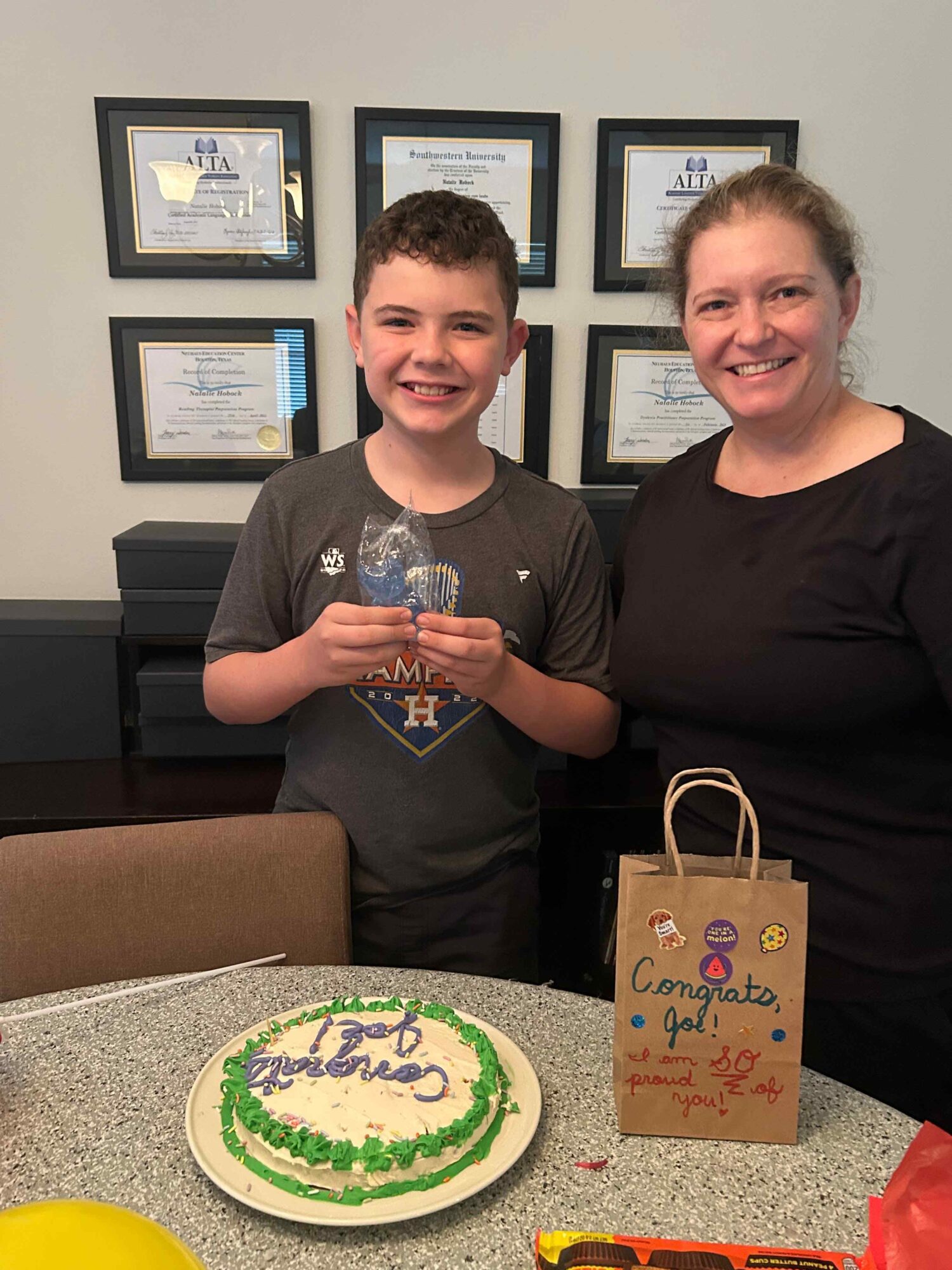 A boy and woman smiling, holding a cake with green and purple decorations, in a room with framed certificates on the wall.
