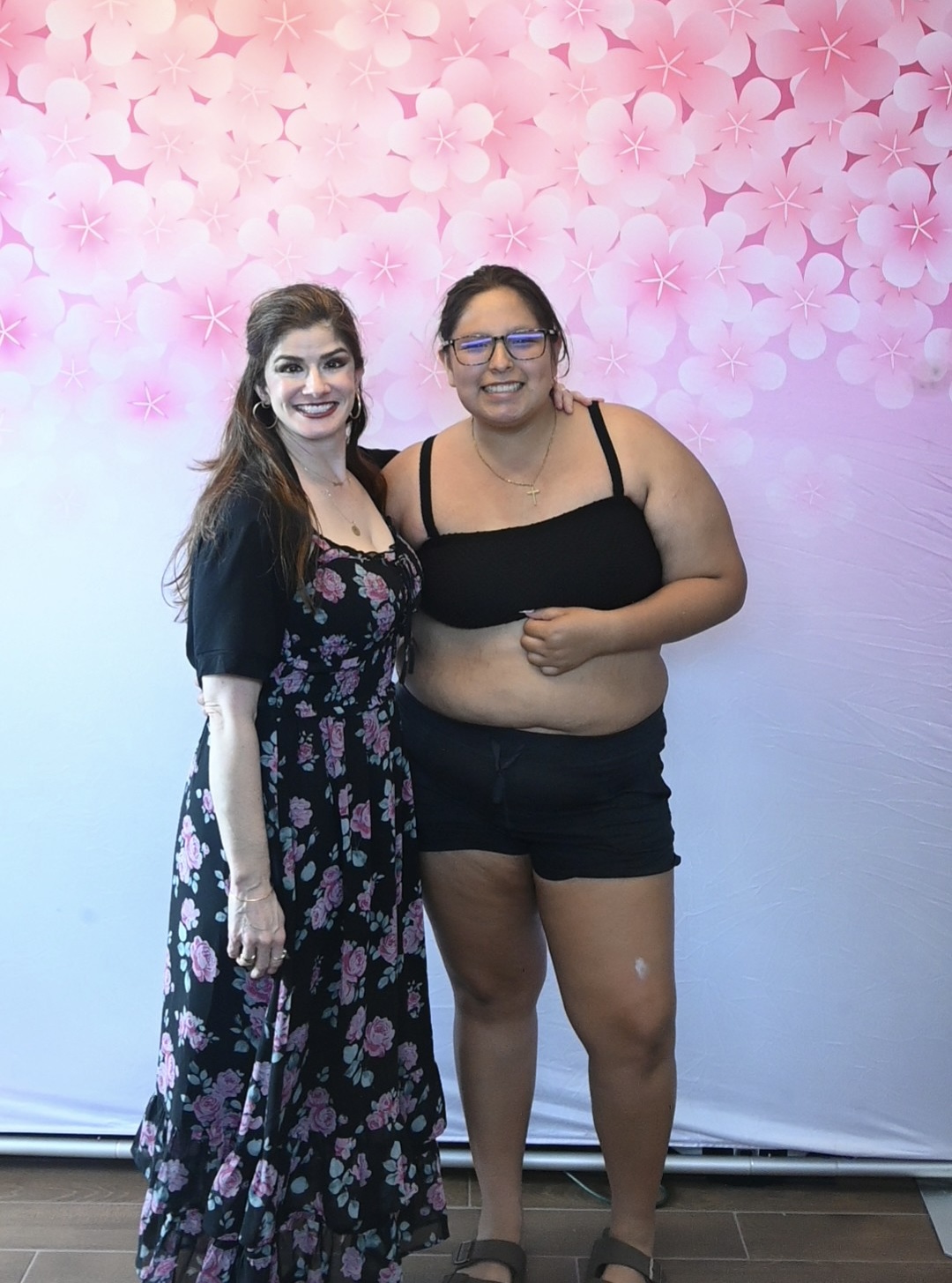 Two women standing together, smiling, in front of a pink floral background, one in a floral dress and the other in a black top and shorts.