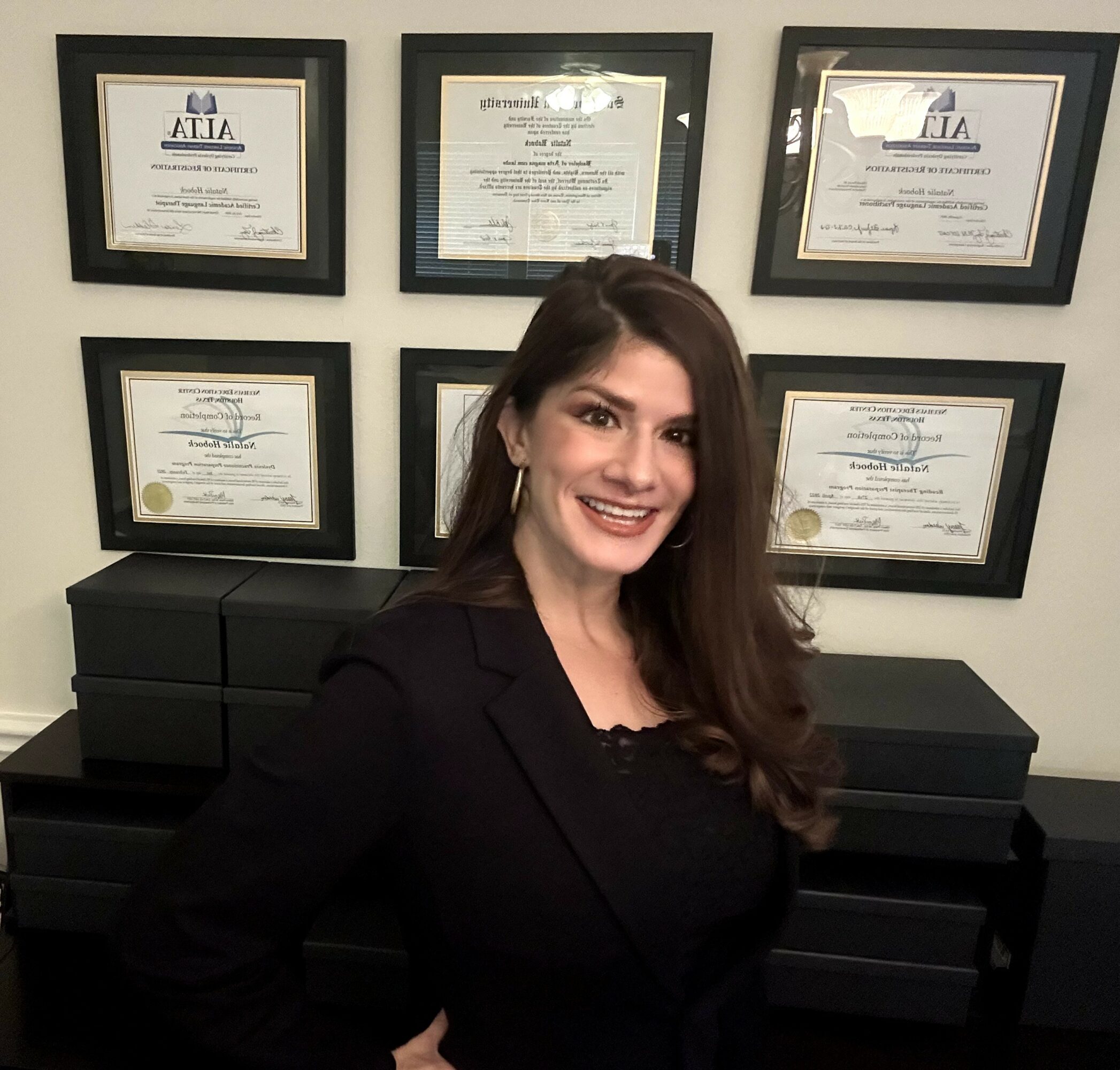 Woman with long dark hair smiling, wearing a black blazer, standing in front of framed certificates on a wall.