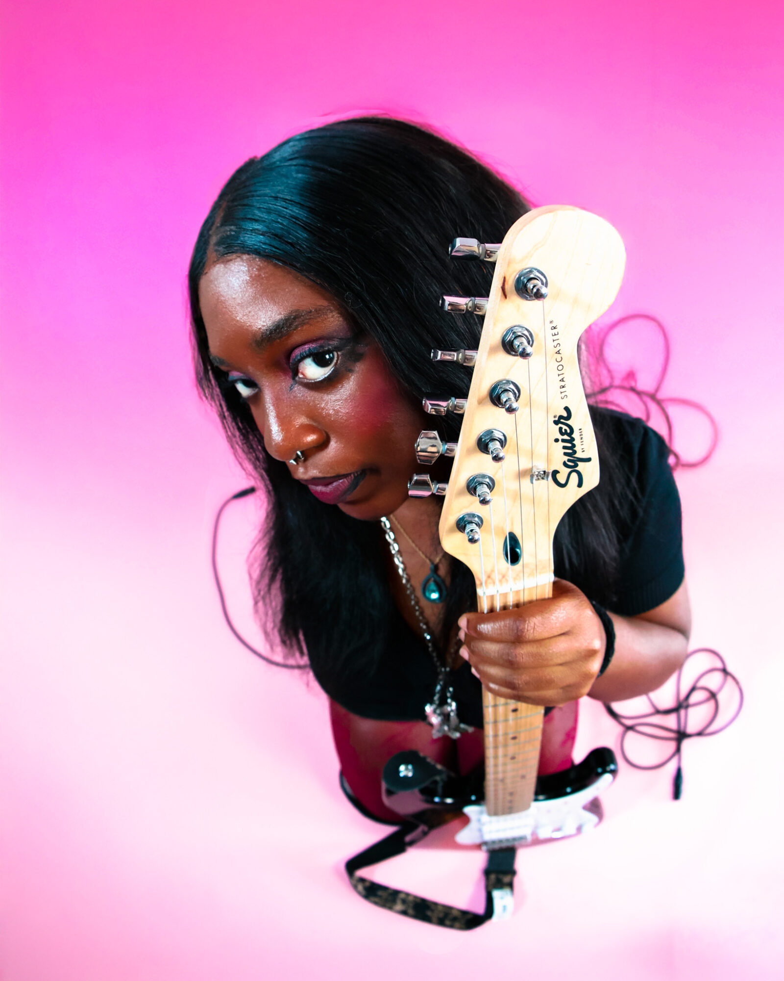 Person with long black hair holding a guitar headstock, against a pink background.