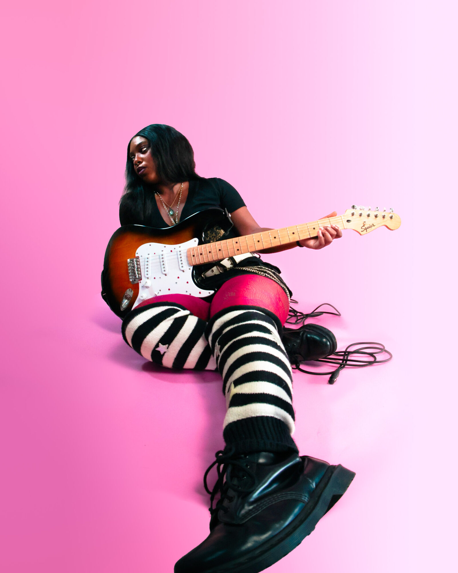 Person sitting on pink floor holding an electric guitar, wearing striped pants and black boots.