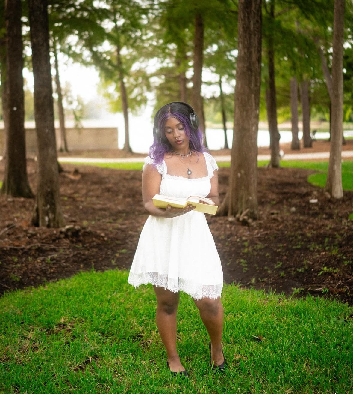 Young woman with purple hair in a white dress reading a book outdoors in a park with trees and grass