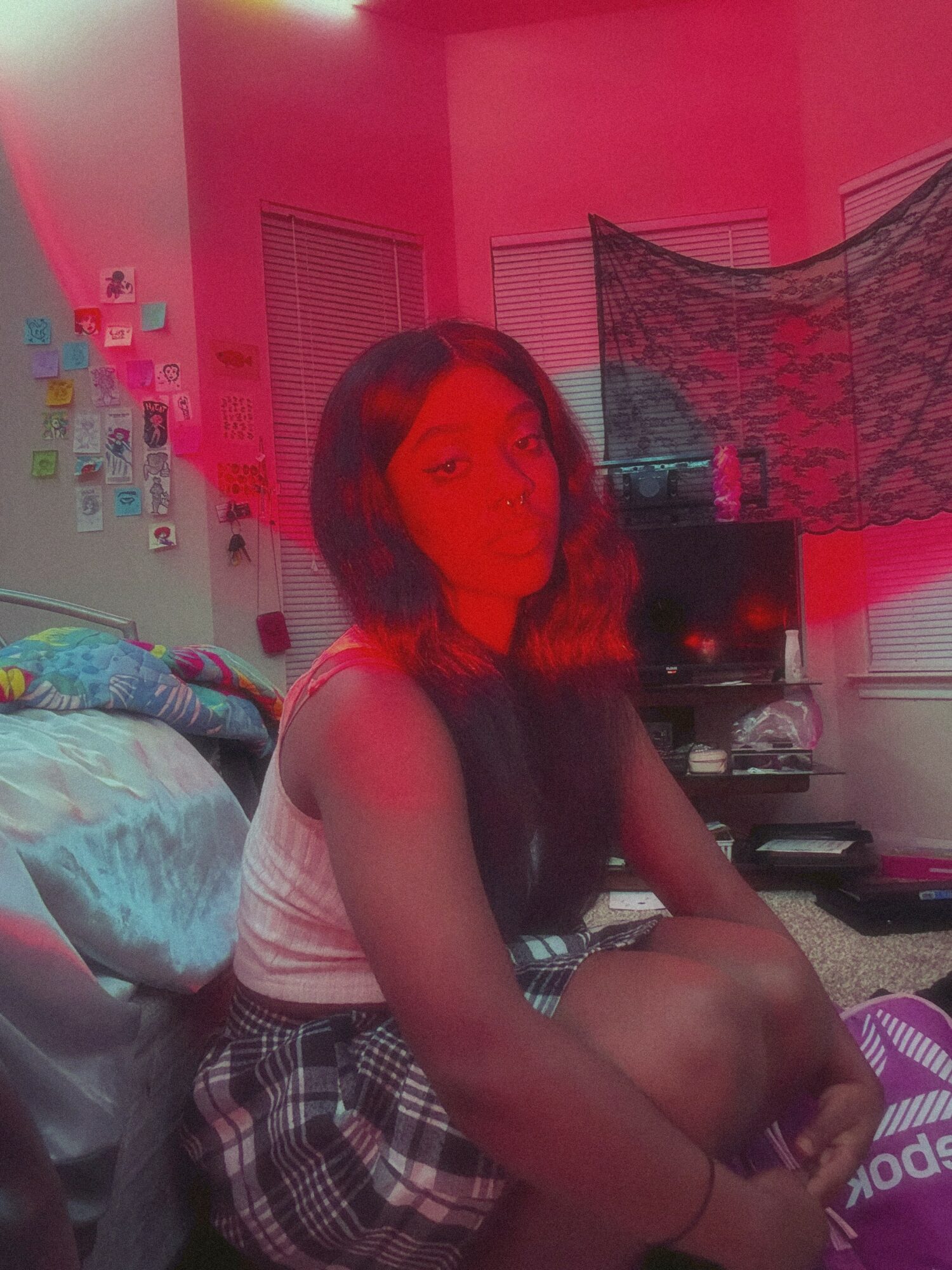 Young woman with long dark hair sitting on a bed in a room with pink lighting, TV, and window blinds.