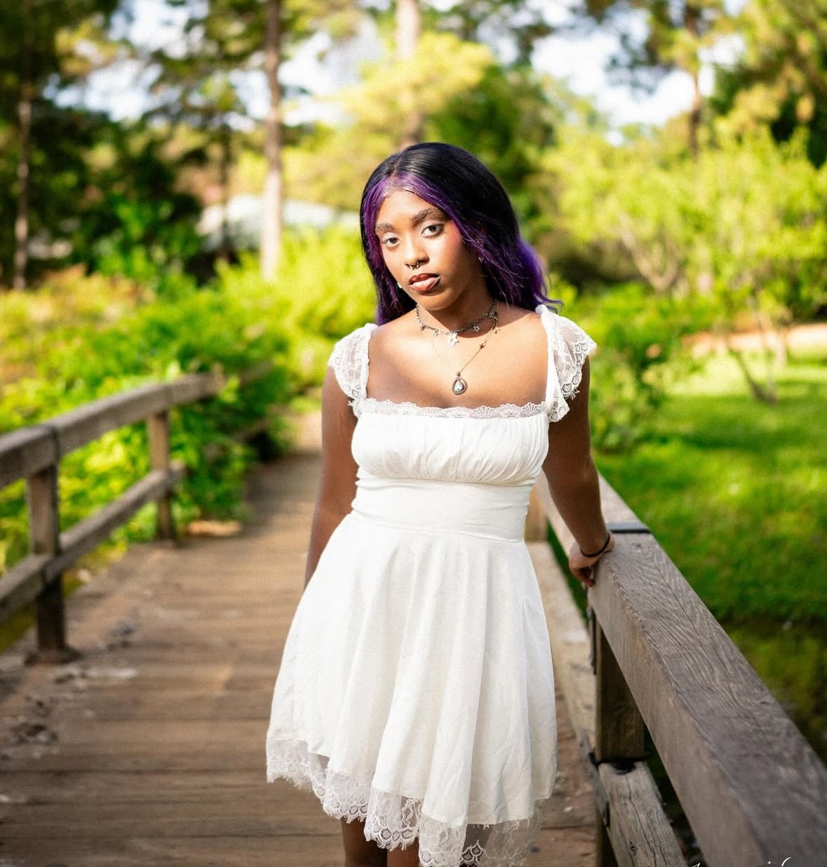 Young woman with purple hair in a white dress standing on a wooden bridge outdoors, surrounded by greenery.