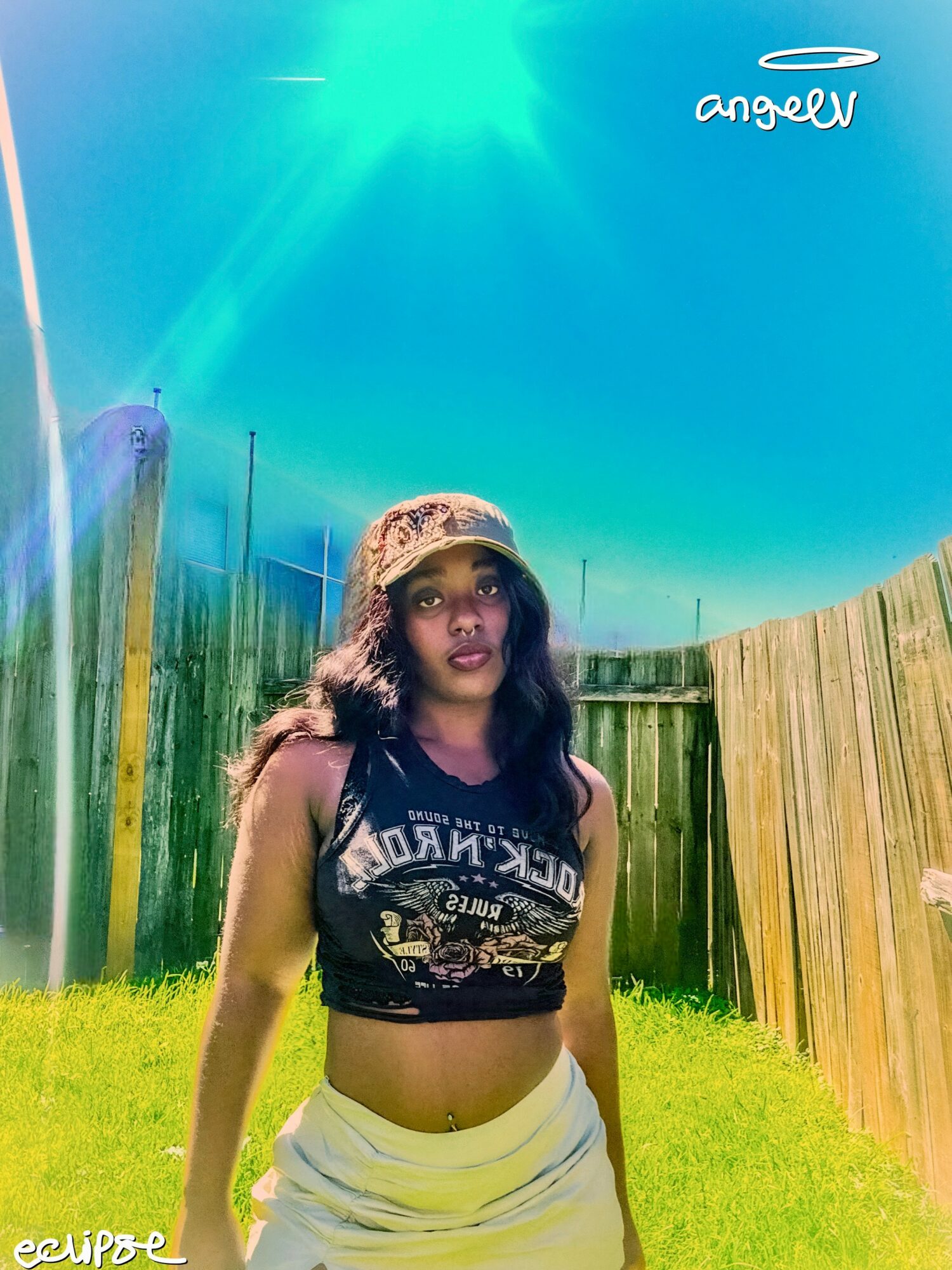 Young woman standing outdoors in a grassy area with wooden fences, wearing a crop top and a hat, under a bright blue sky.