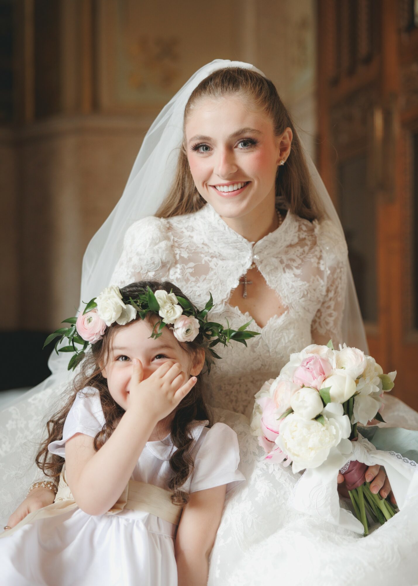 A bride and a young girl, both smiling, with the bride holding a bouquet of flowers, in an indoor setting.