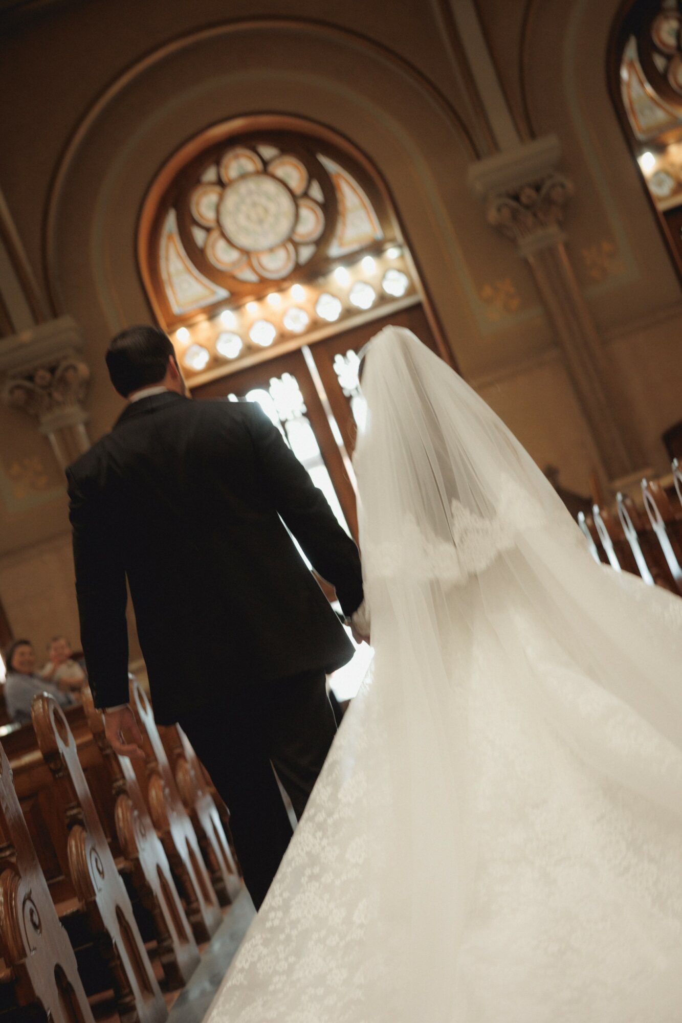 A bride and groom walk down the aisle in a church with stained glass windows, holding hands, with rows of chairs on sides.